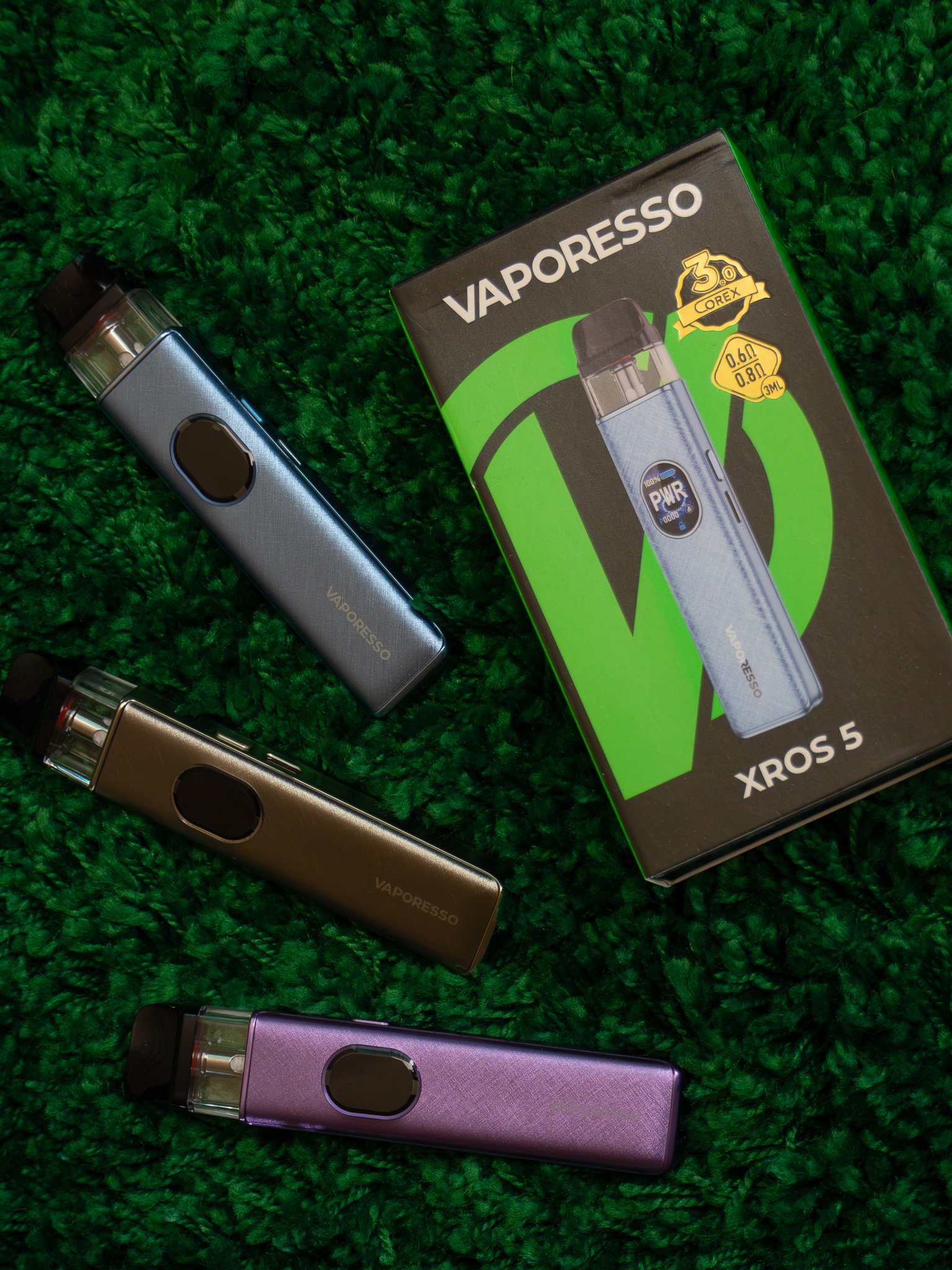 Colorful Vaporesso Xros 5 vaping devices in purple, gold, blue, and black colors placed on a green textured surface along with their packaging.