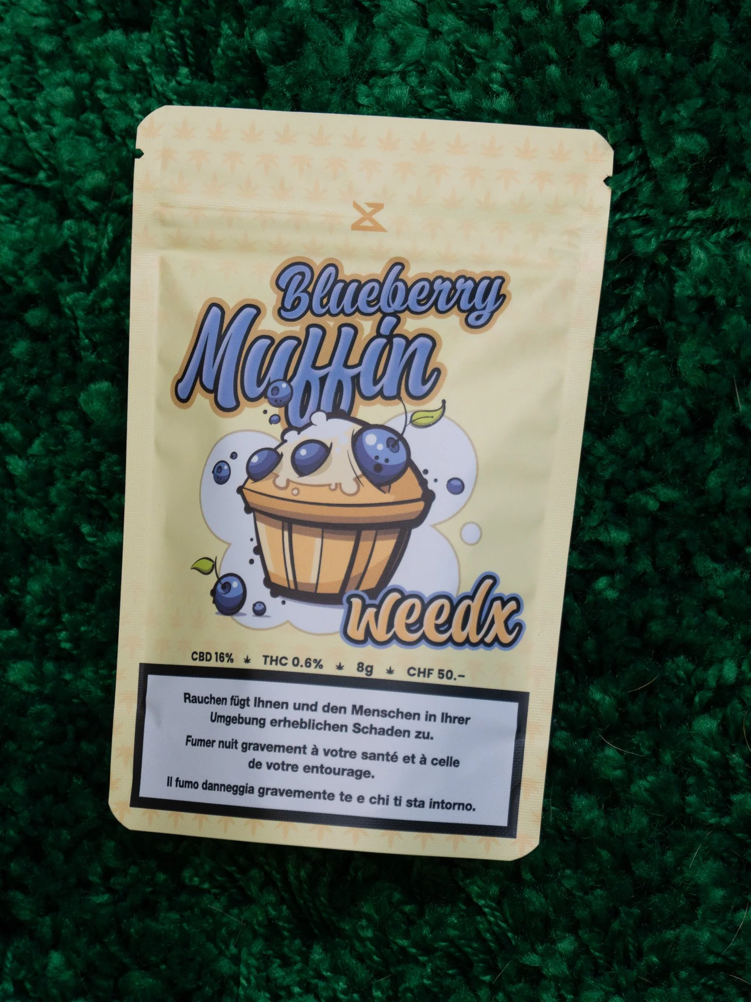 Packaging of blueberry muffin with cartoon muffin illustration and blueberries on it, placed on a green textured surface.