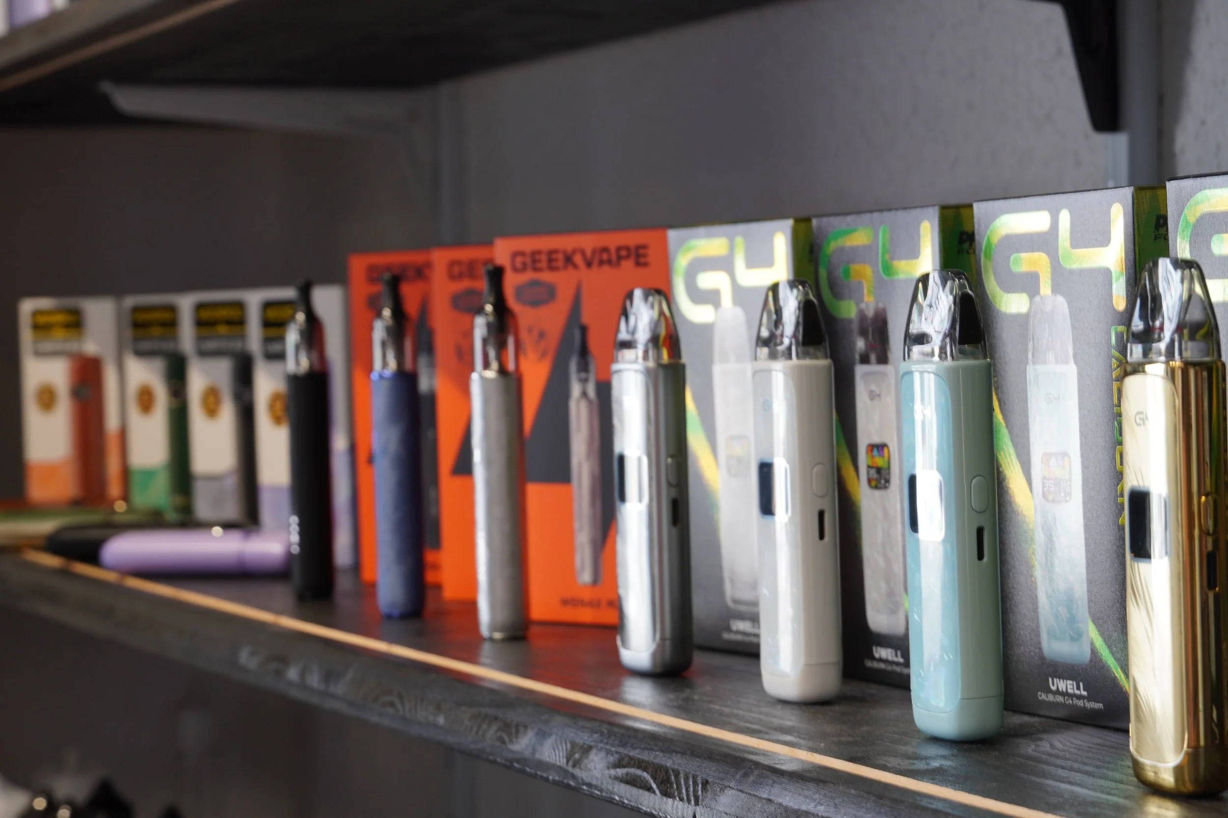 A shelf displaying various vape devices and e-liquid boxes, including GeekVape and Caliburn models.