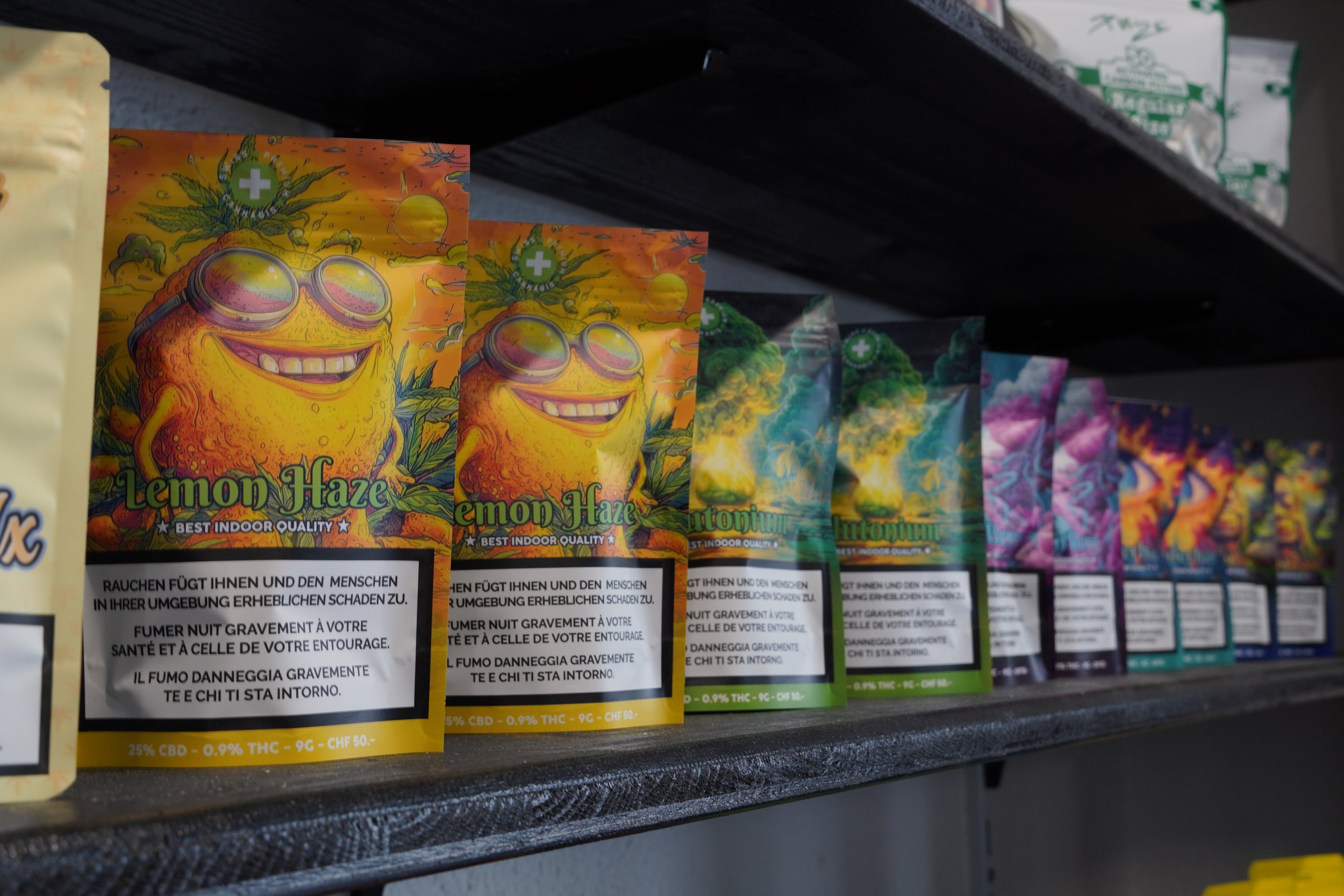 Colorful cannabis product packages on a store shelf, labeled Lemon Haze and other strains, with warning labels in multiple languages.