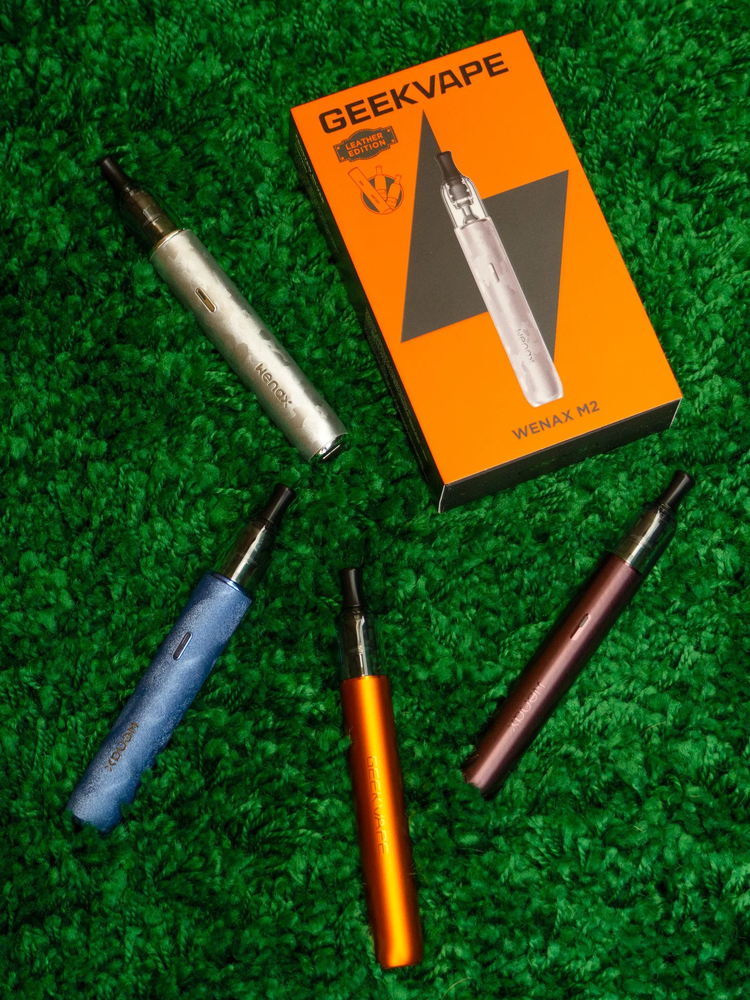 Multiple colorful vape pens of various colors, including blue, orange, and rose gold, lying on a green textured surface. A box labeled 'GEEKVAPE' and 'WENAX M2' is also visible.
