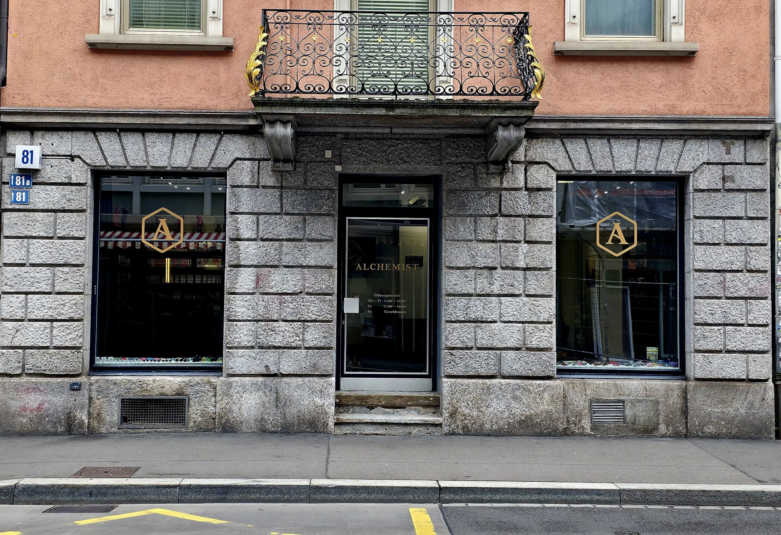 Storefront with a sign that says 'ALCHEMIST', two large display windows with a gold 'A' logo, a glass door with business hours, and a small balcony with decorative iron railing above the entrance.