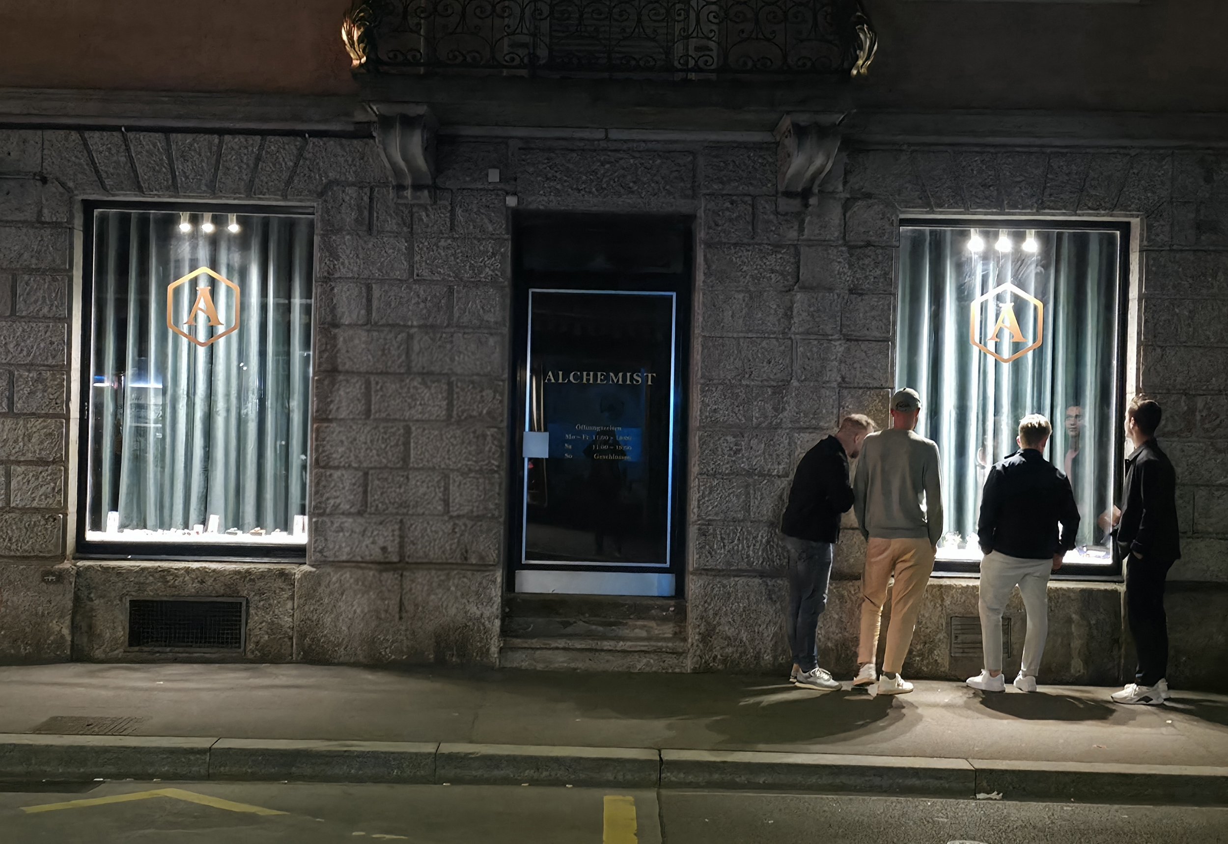 Nighttime exterior of a stone building with two large display windows and a central door, advertising 'ALCHEMIST.' Several people are standing and conversing in front of the entrance.