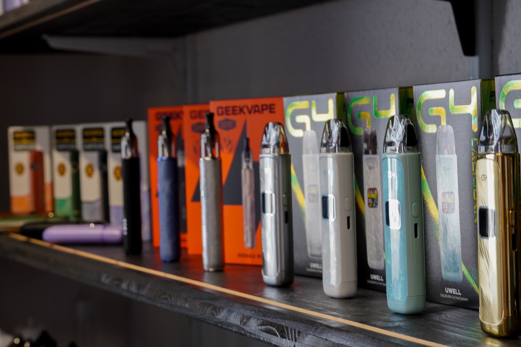 A store shelf with various vaping products, including multiple vape pens and e-cigarette bottles, displayed in packaging and boxes.