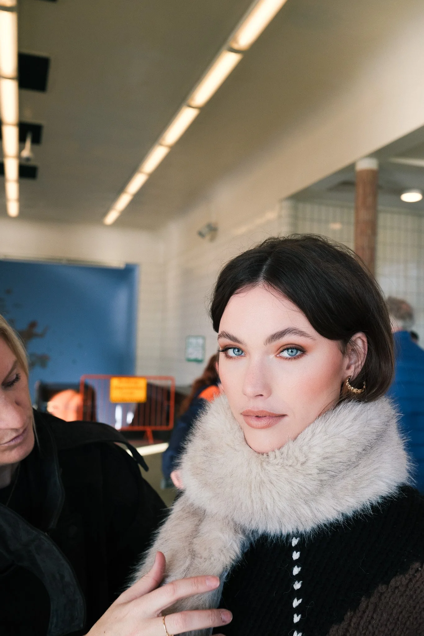 A woman with blue eyes and dark hair wearing a large white fur collar, looking at the camera in an indoor setting with fluorescent lighting.