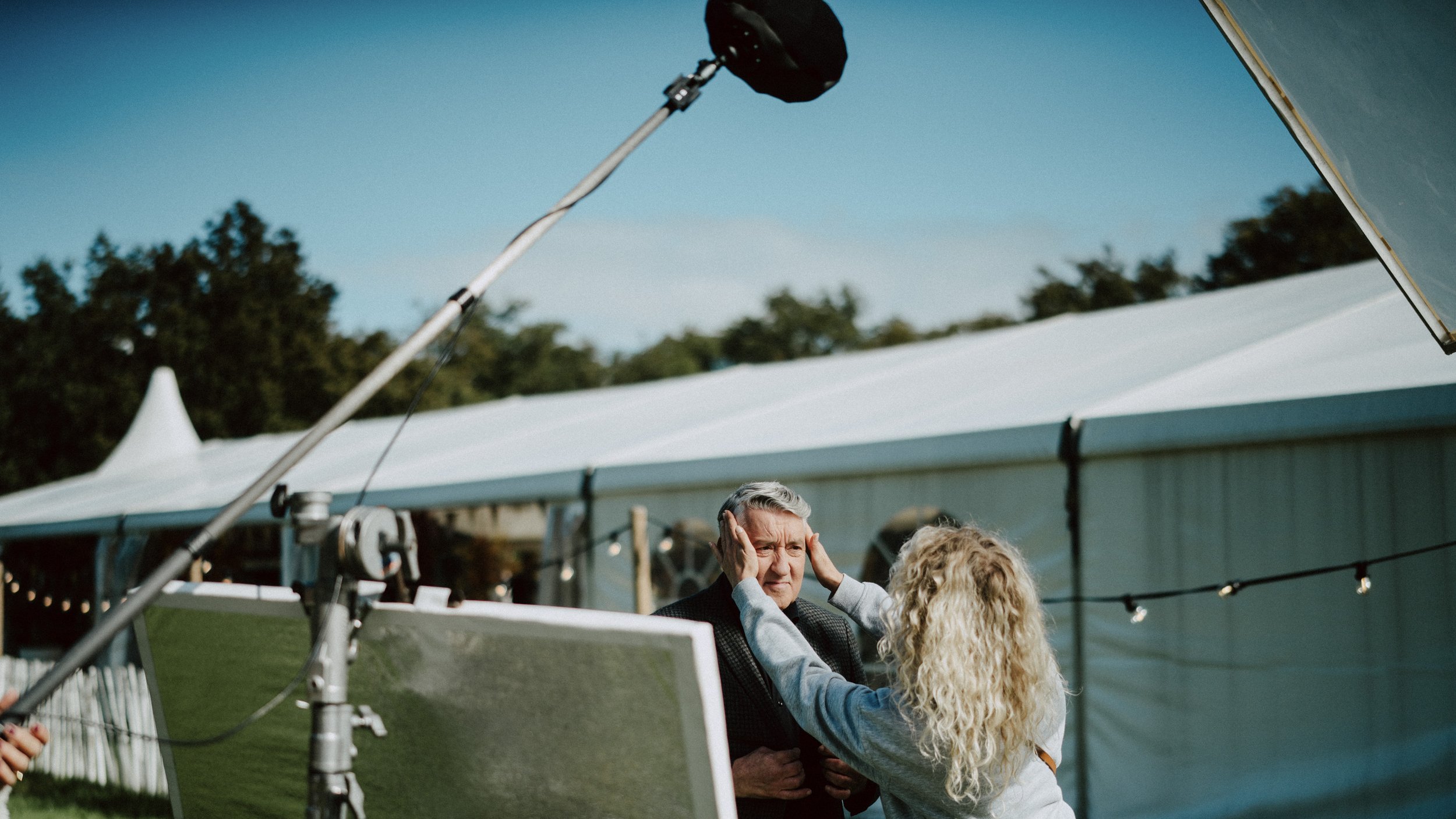 A man with gray hair and a woman with curly blonde hair are standing outdoors at an event, with a large white tent and trees in the background. The woman is touching the man's face as they converse.
