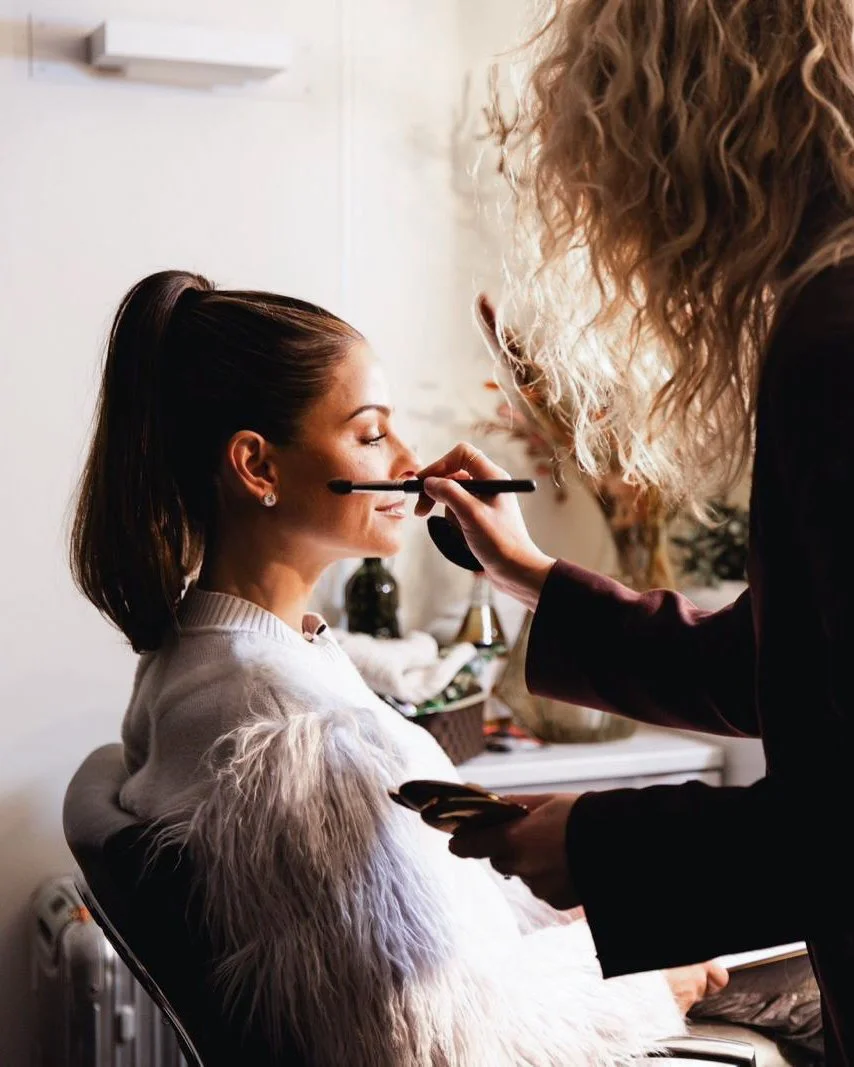 A woman with dark brown hair in a high ponytail is getting her makeup done by a makeup artist with curly blonde hair. The woman is sitting with her eyes closed, wearing a white sweater with fluffy sleeves, and appears relaxed.
