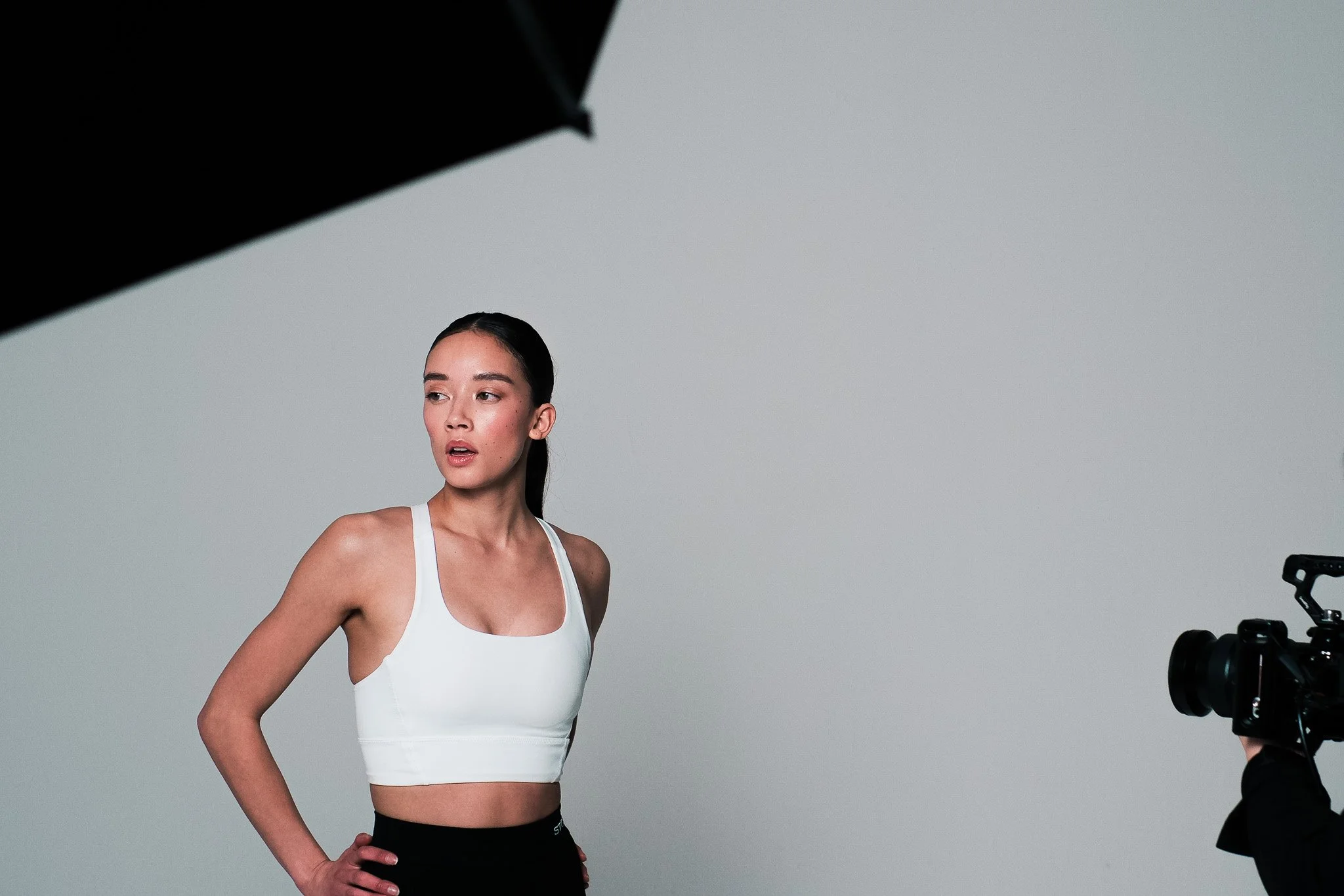 A woman with dark hair pulled back, wearing a white sports bra and black workout leggings, is standing with one hand on her hip and looking slightly to the side during a photo shoot in a studio with a gray background. A camera and lighting equipment 