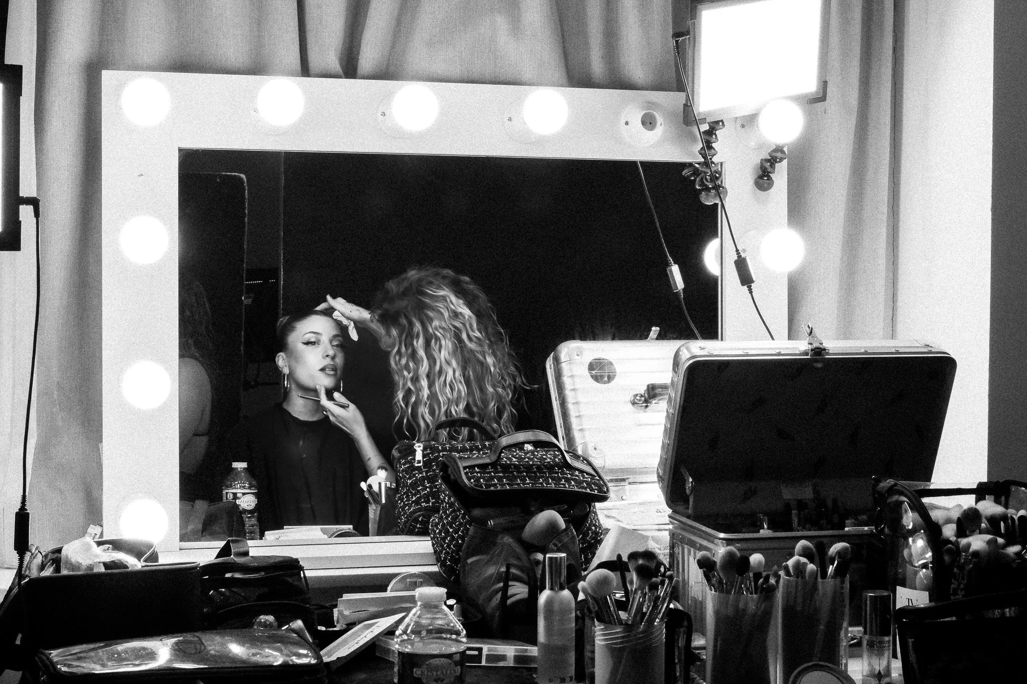 A woman with makeup brushes applying makeup to a client in front of a mirror with light bulbs.