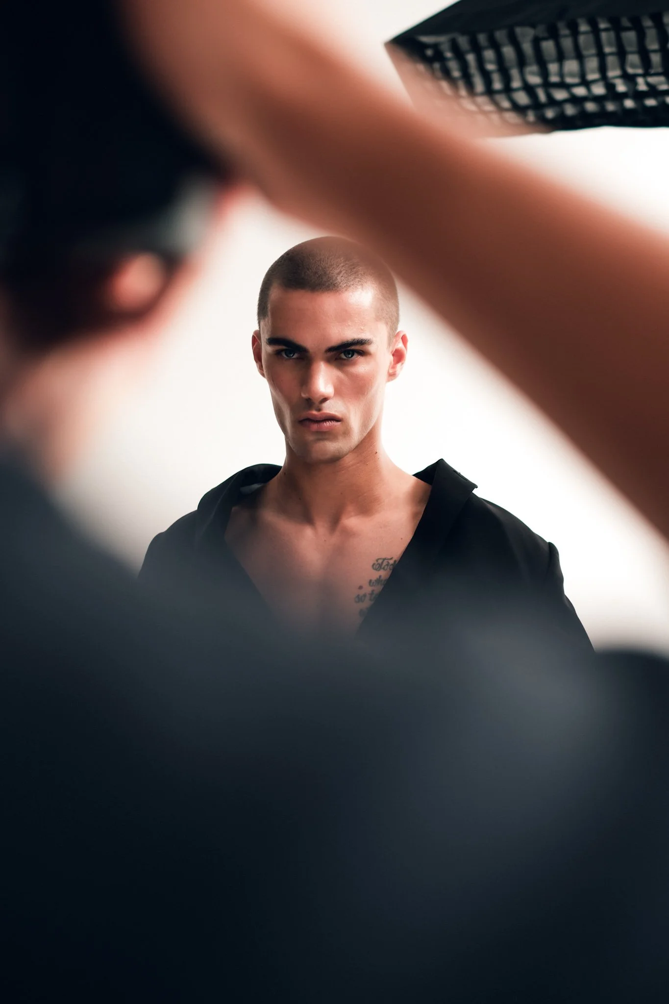 A young man with a shaved head and tattoos on his chest looking directly into the camera, taken from an unusual perspective with blurred objects framing the image.
