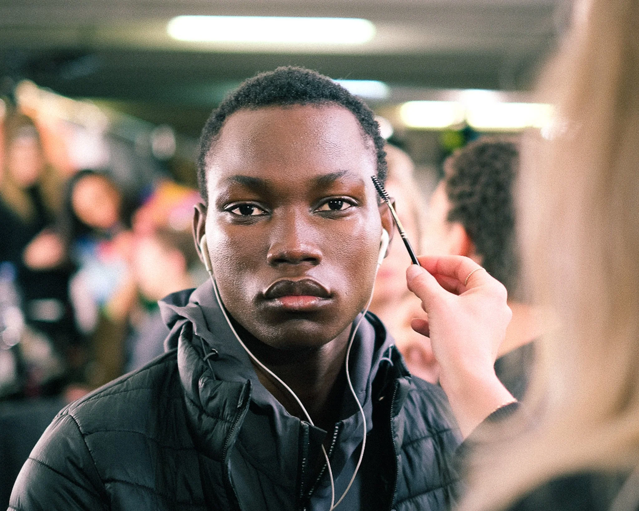 A young woman with short, curly hair is smiling and looking directly at the camera while applying eye makeup with a mascara wand. She is wearing earphones and a black jacket, and is in a busy indoor setting with other people in the background.