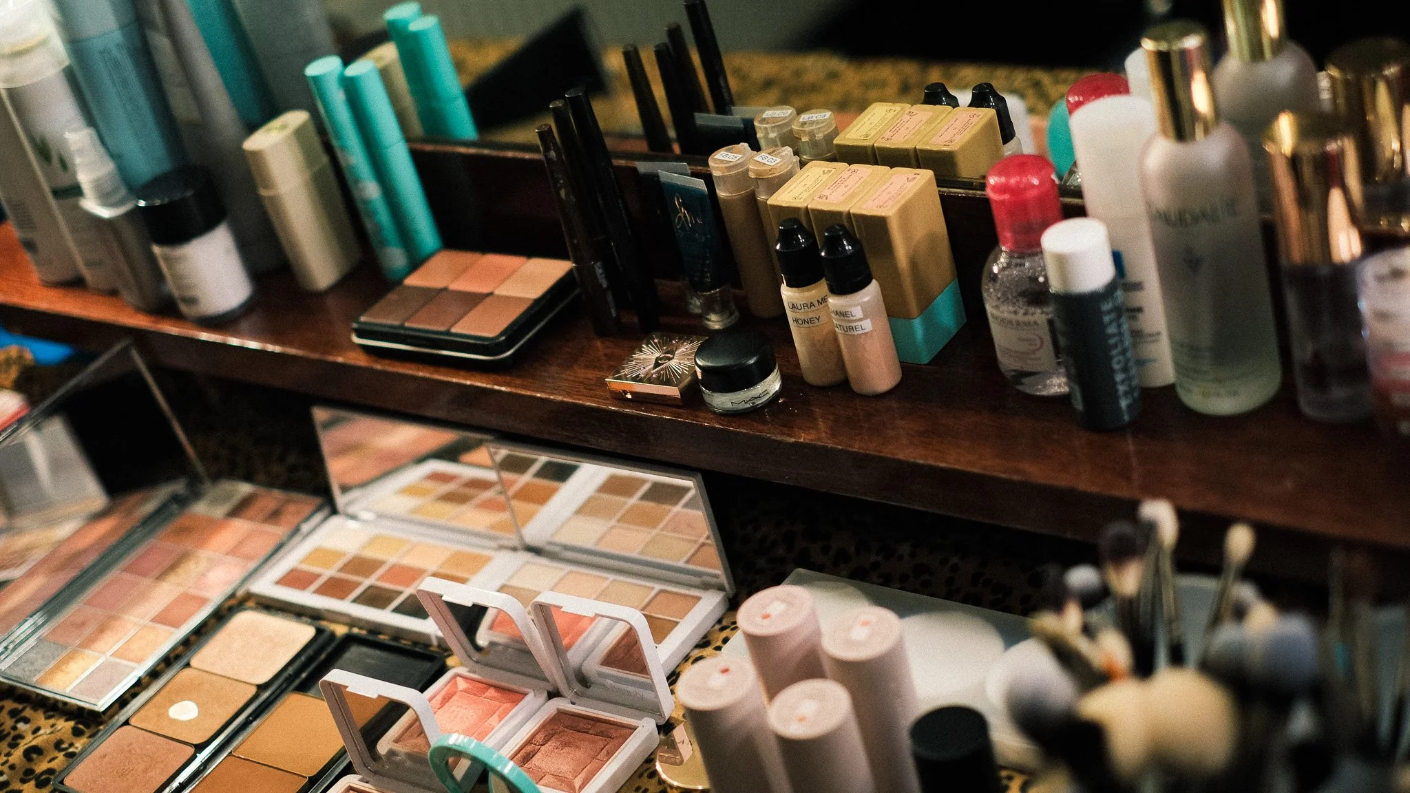 A collection of makeup products including powders, sprays, creams, and brushes organized on a wooden surface with a mirror in the background and open makeup palettes in the foreground.