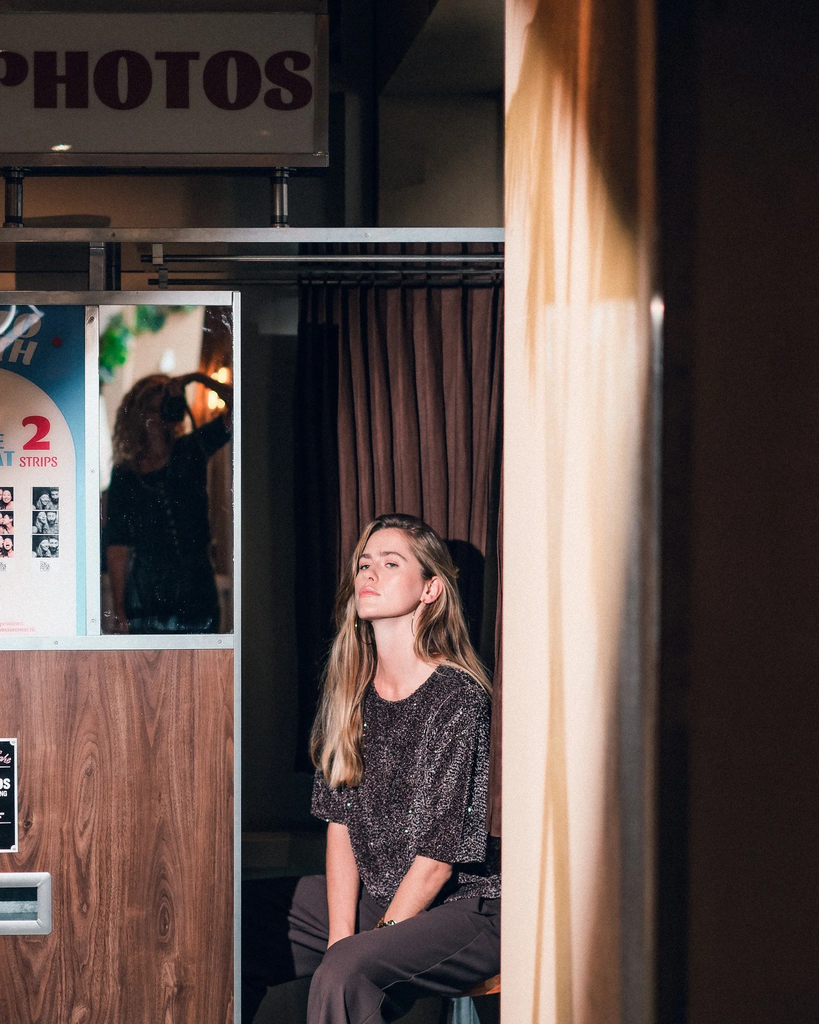 A woman with long blonde hair, wearing a shiny black top, sitting on a wooden stool in a photo booth with a curtain behind her. There is a sign above that says 'PHOTOS' and a partially visible photo strip on the left side of the image.