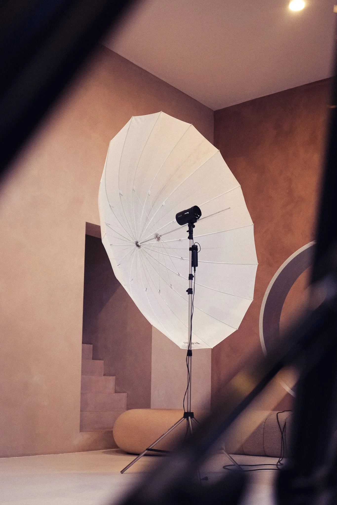 A photography lighting setup in an indoor space featuring a large white umbrella reflector on a stand, with a black studio light attached, positioned in front of a beige wall with stairs and a circular architectural feature visible in the background.