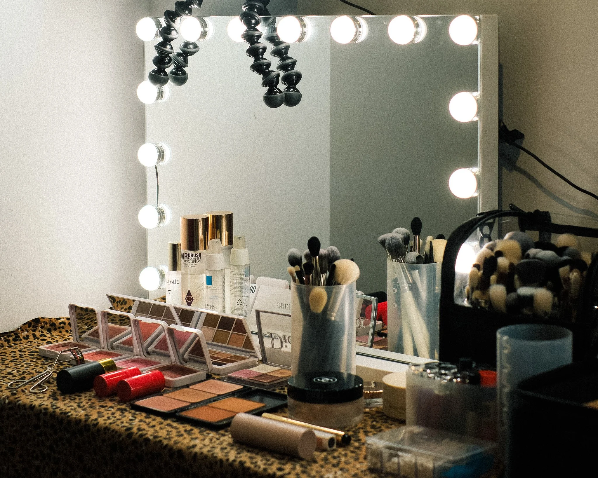 A makeup vanity station with a mirror framed by round lights, various beauty products including makeup brushes, foundation bottles, an eyeshadow palette, and makeup tools on a leopard-print cloth, and some bottles and containers on the countertop.