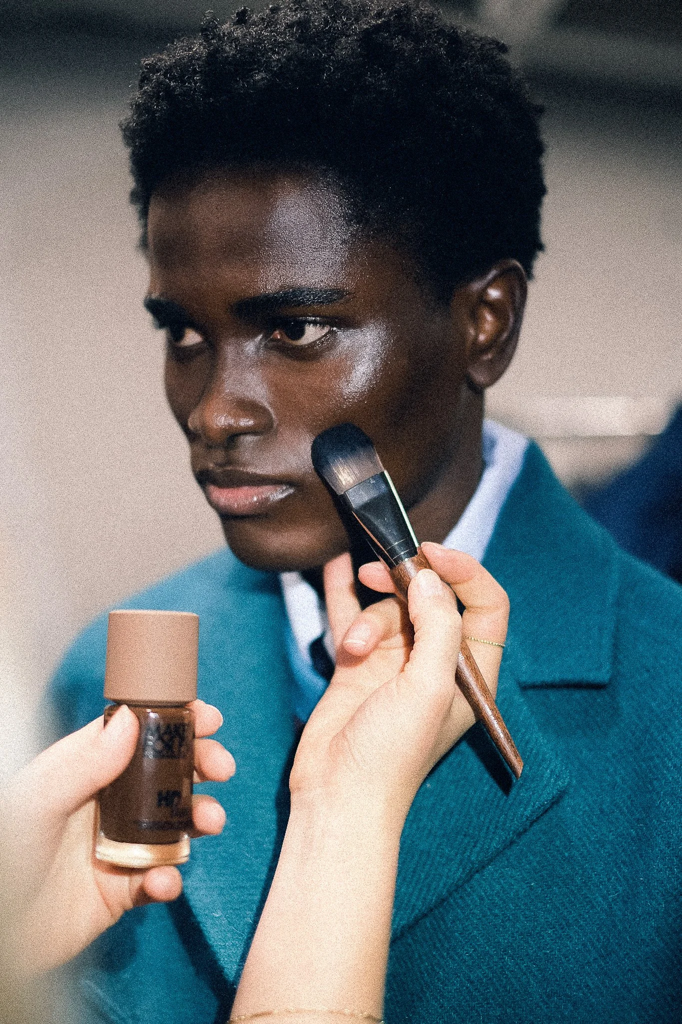 A makeup artist applying foundation to a young woman with dark skin and short curly hair, using a brush and small container of makeup.