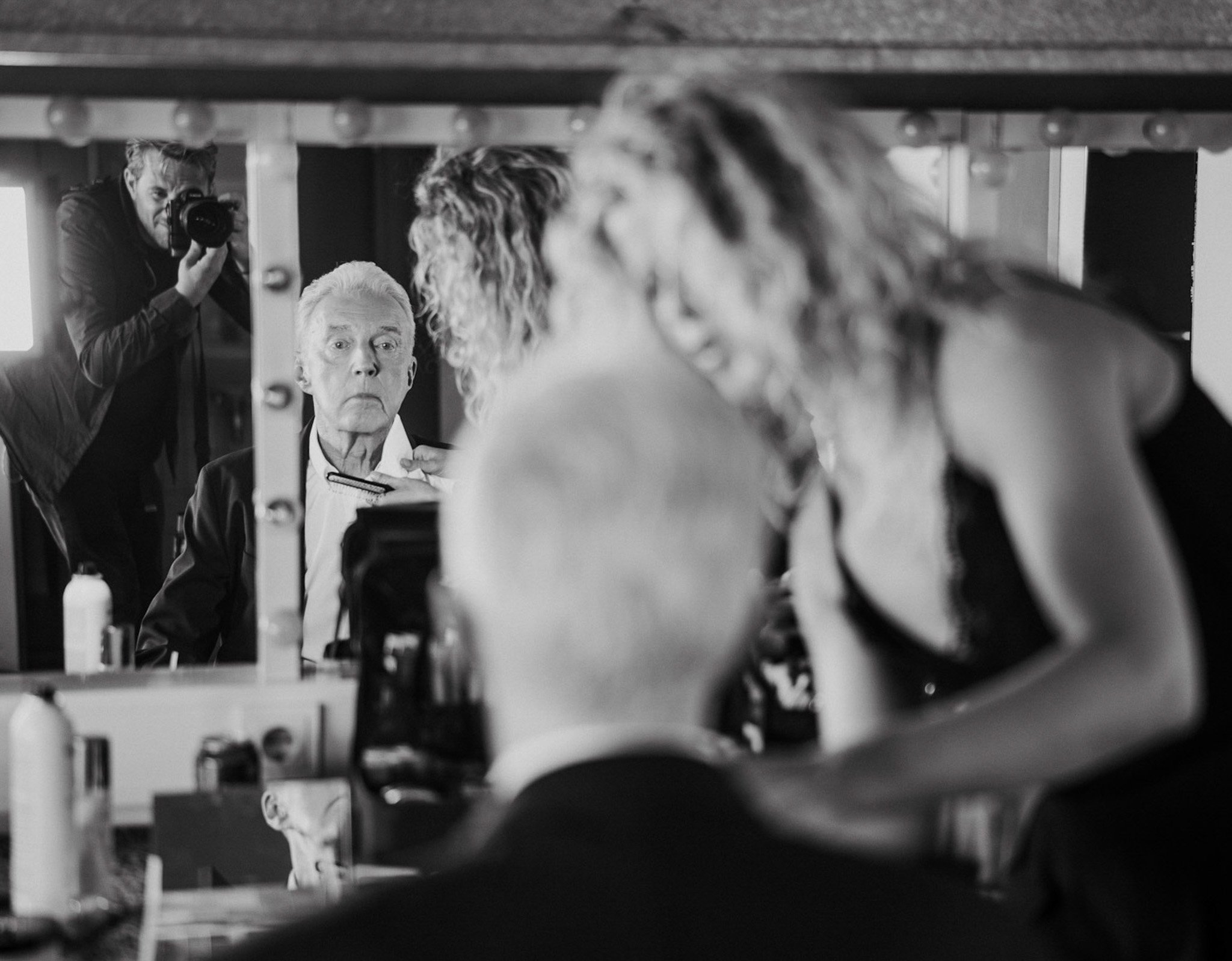 Black and white photo of a woman getting her hair styled by a hairstylist, with a man taking a photograph in the background.