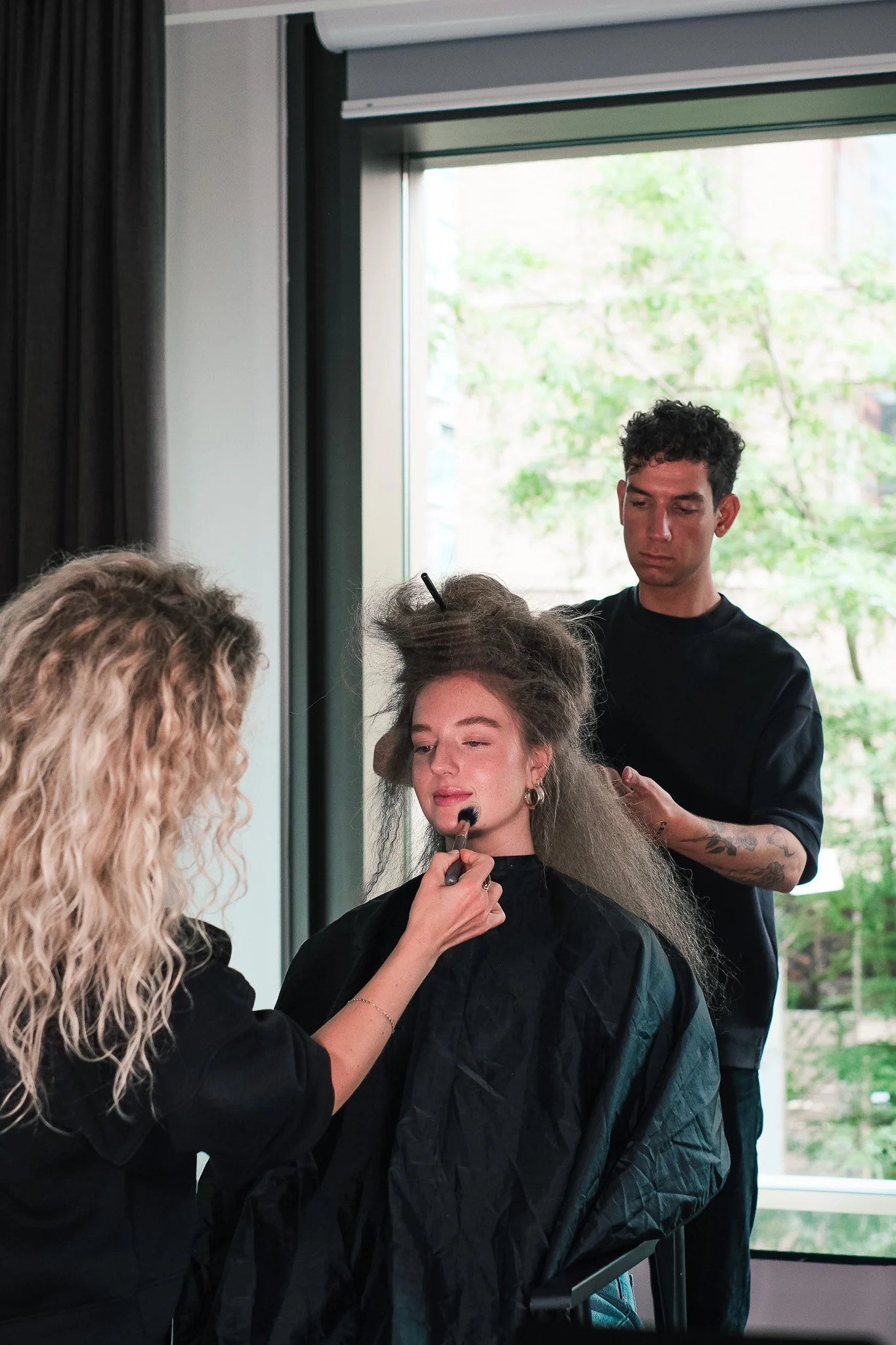 A woman with curly blonde hair applies makeup to a woman with long dark hair seated in front of a mirror, while a man with short curly black hair, tattoos, and a black shirt looks on.