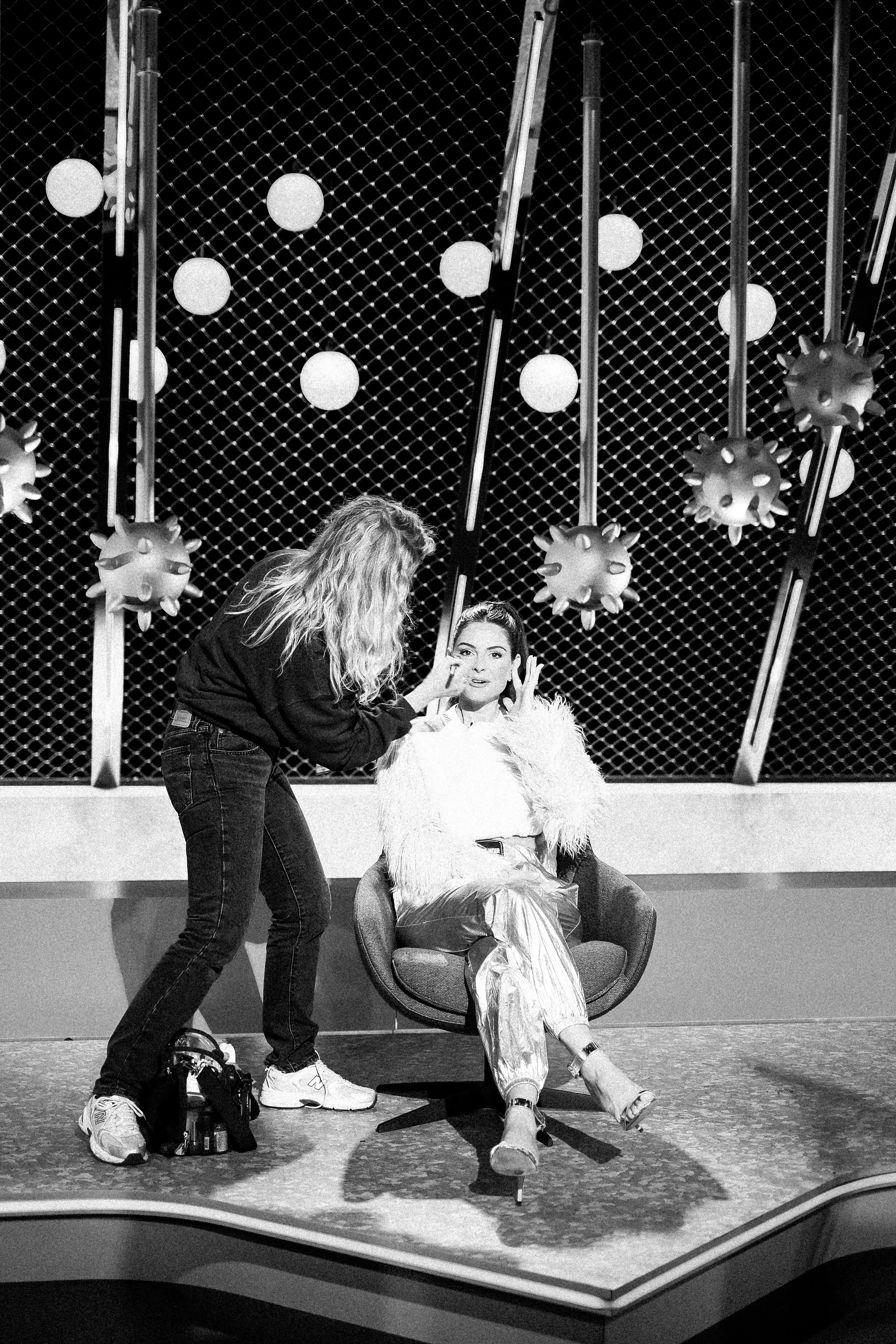 A woman with long hair and a woman with a high ponytail sit in a chair, while another woman with curly hair applies makeup or makeup tools to the woman with the high ponytail. They are on a stage with a patterned backdrop with hanging decorations.