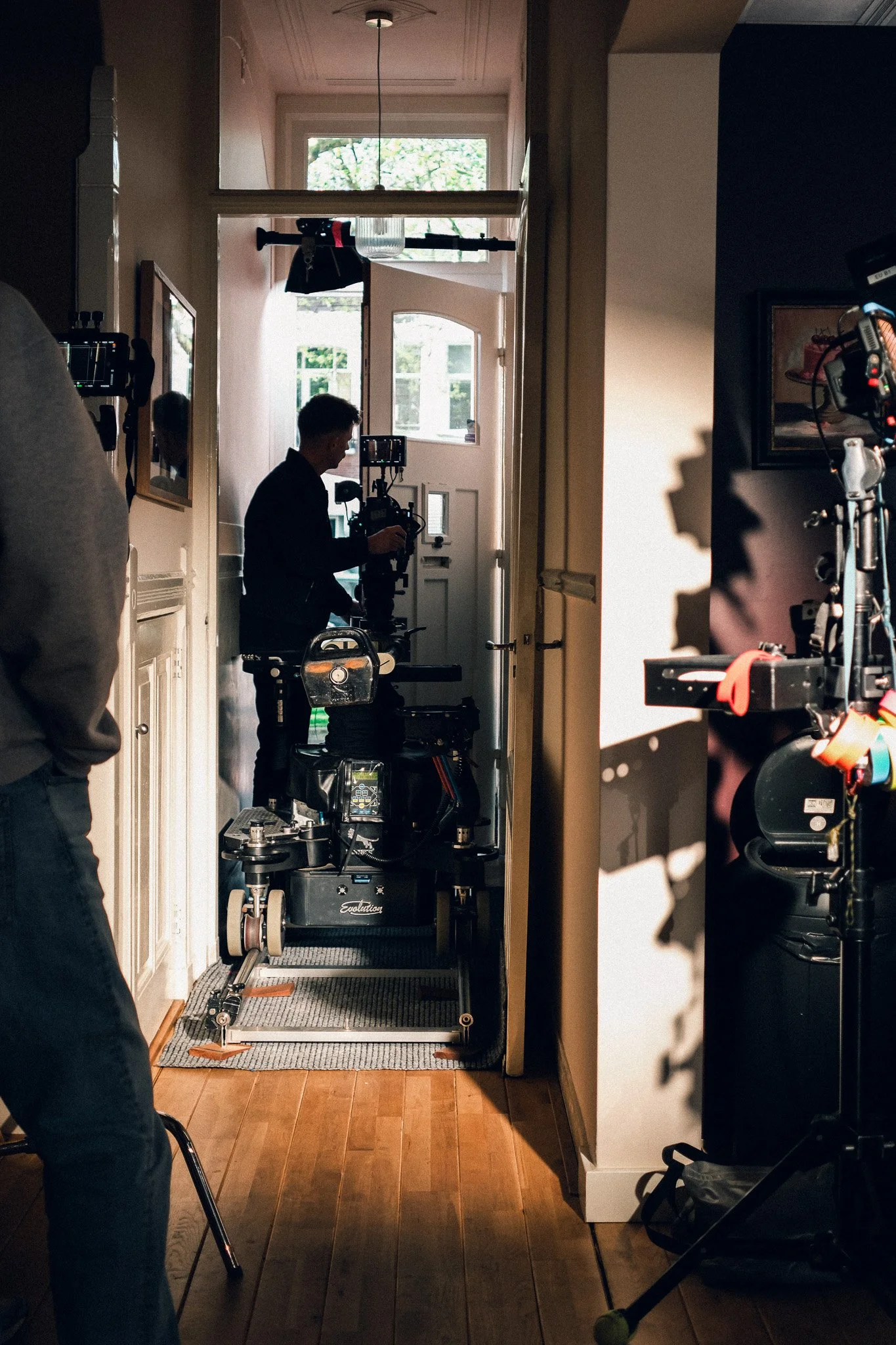 A film crew setting up equipment in a narrow hallway of a house. A person operating a camera on a cart, with lighting and other filming gear around. Sunlight is coming through the open front door and windows.