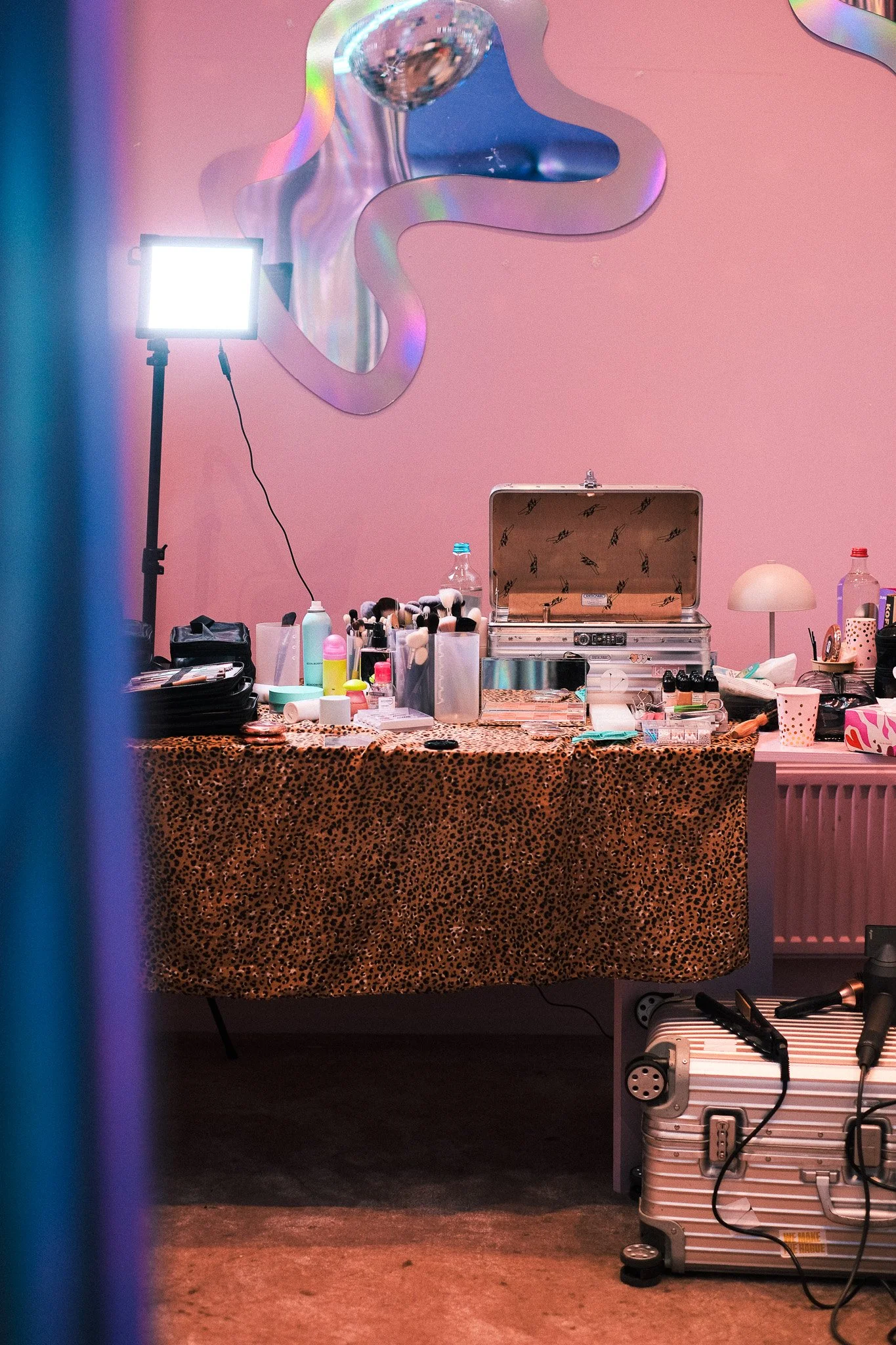 A makeup station with various cosmetics, brushes, and tools on a leopard print tablecloth, a lunch box, and a water bottle. A bright light is on a tripod against a pink wall with a metallic, looping decorative piece. There is a small white table with