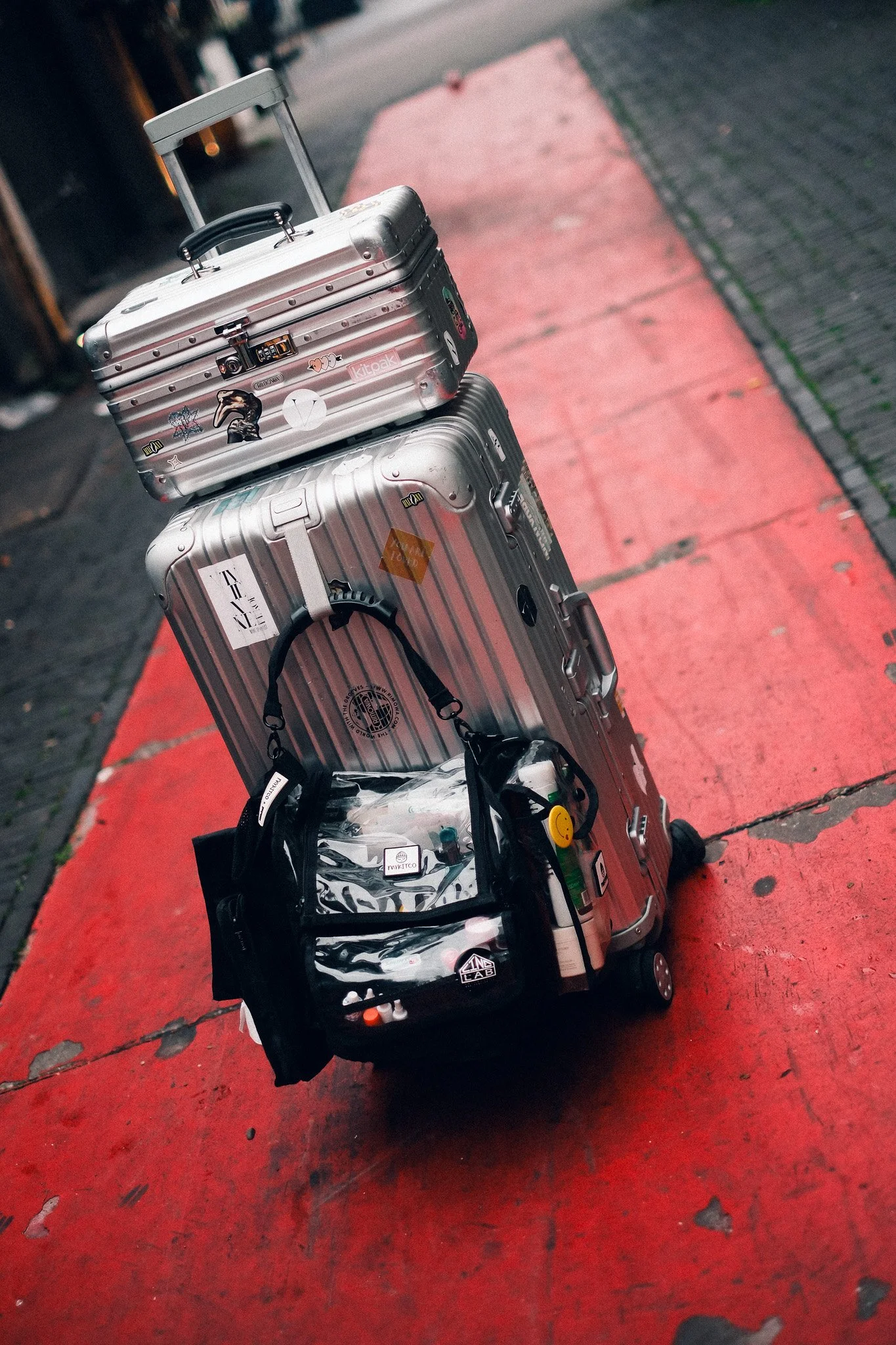 Three pieces of rolling luggage and a backpack are stacked on a red sidewalk. The luggage is silver with various stickers and tags, and the backpack is black with a clear pocket containing items inside.