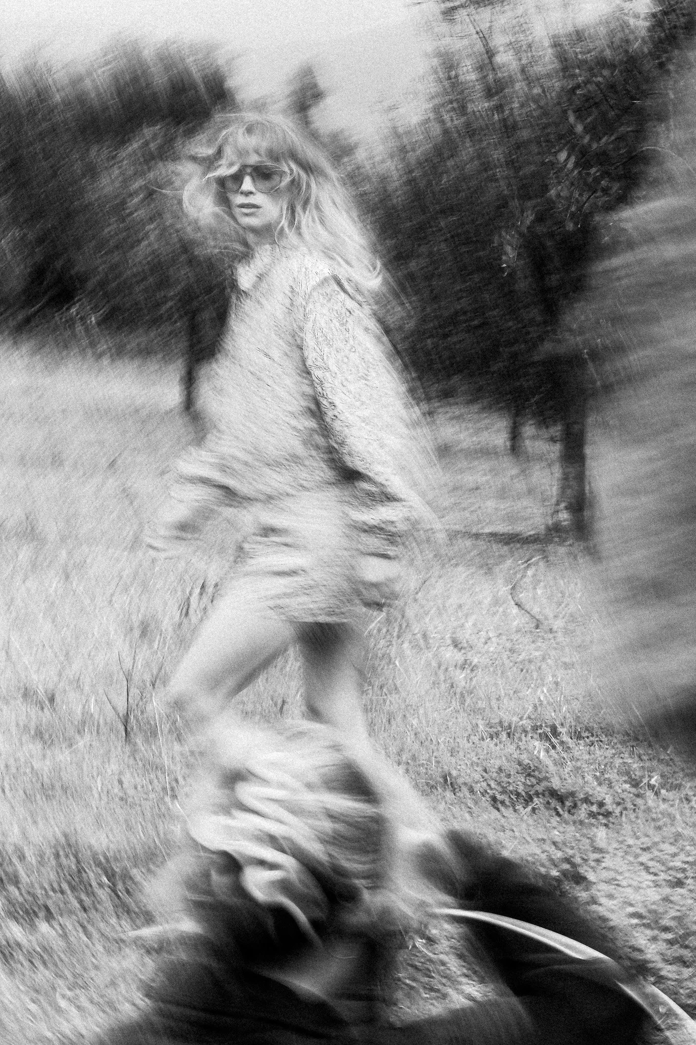 A woman with long wavy hair, wearing sunglasses and a textured coat, walking outdoors on grass with trees in the background. The photo has a blurred, motion effect.