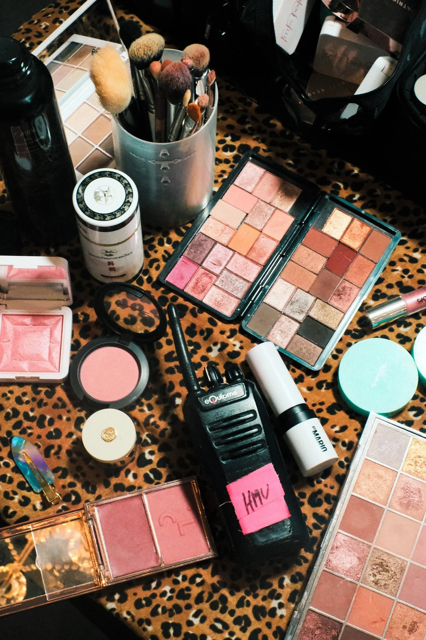 Makeup and beauty products arranged on a leopard print surface, including eyeshadow palettes, blush, brushes, a spray bottle, and various makeup tools.