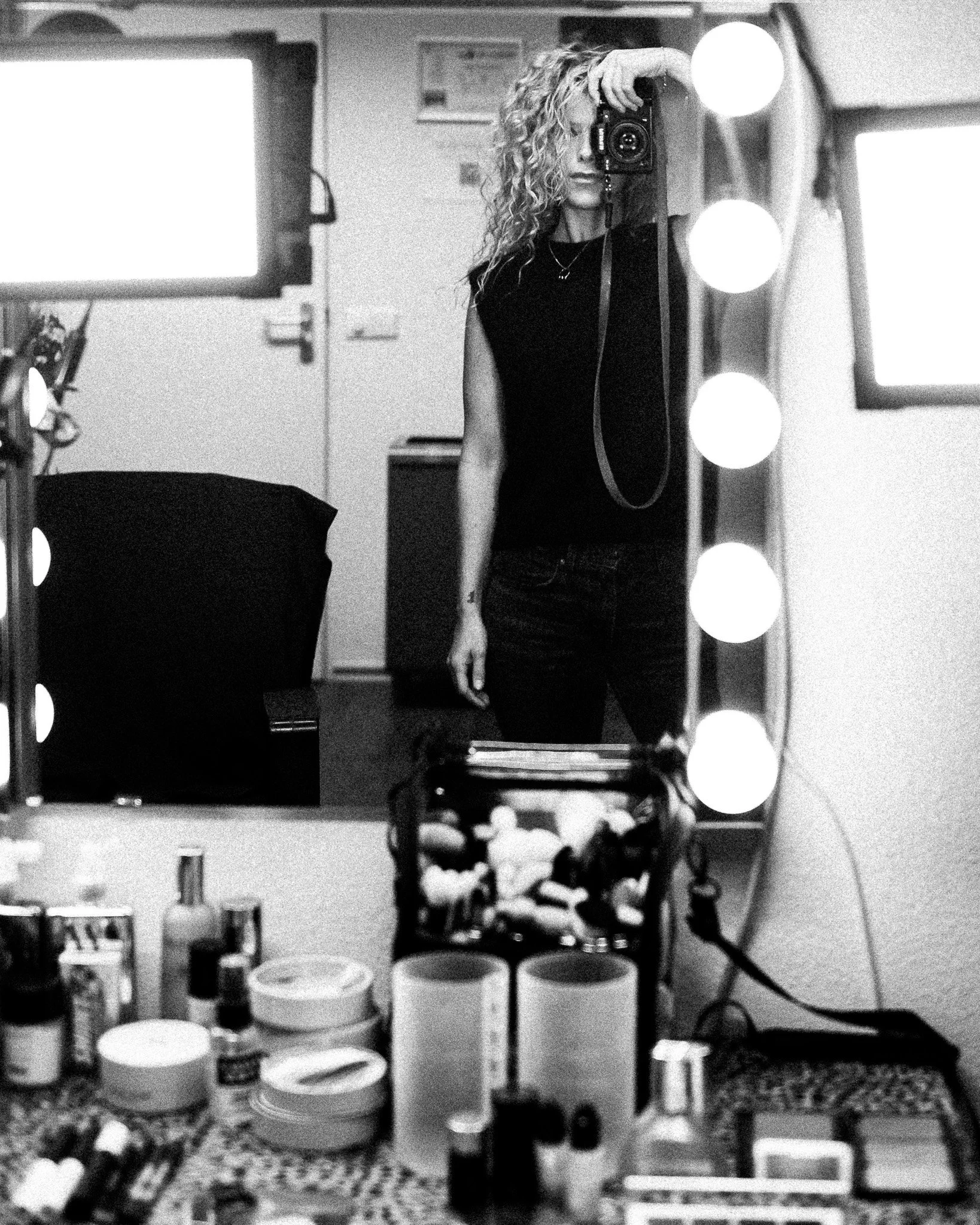 A woman with curly hair taking a selfie in front of a mirror with her camera. The scene is illuminated with large round vanity lights around the mirror. In the background, shelves with various items are visible.
