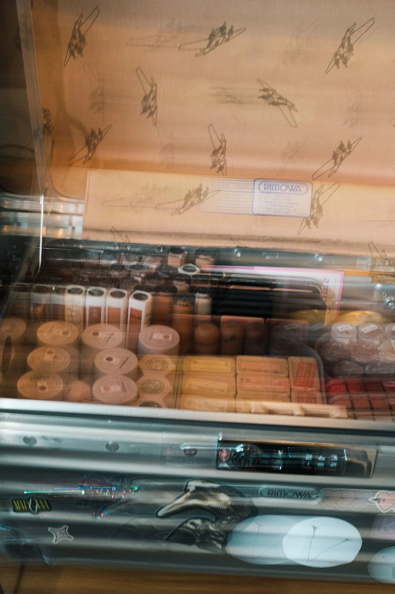 Photo of a display case filled with various makeup products, including foundation bottles, lipsticks, and other cosmetics, viewed through the glass of the case.