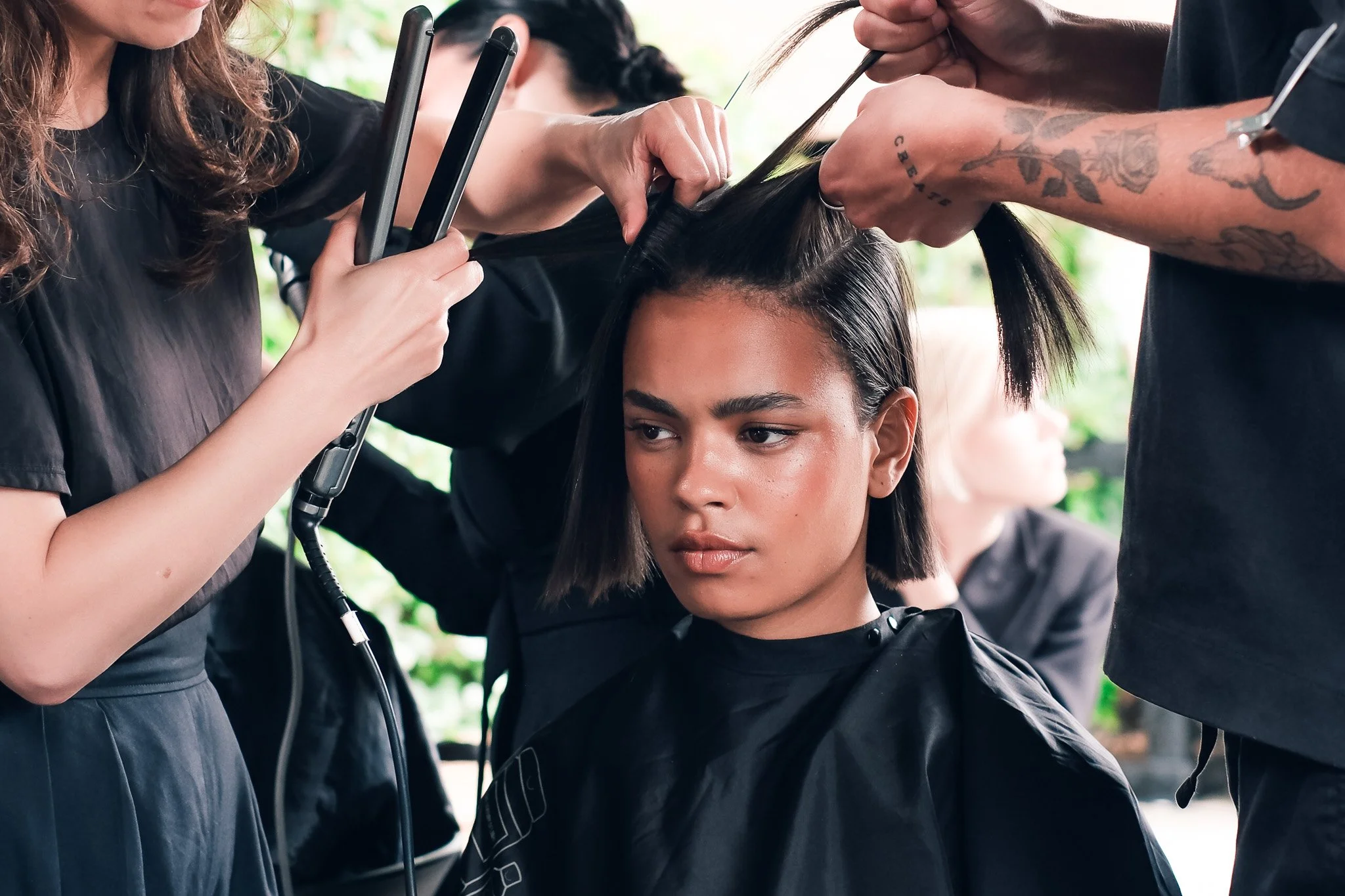 A woman with shoulder-length dark hair is getting a haircut, surrounded by hairstylists working on other clients.