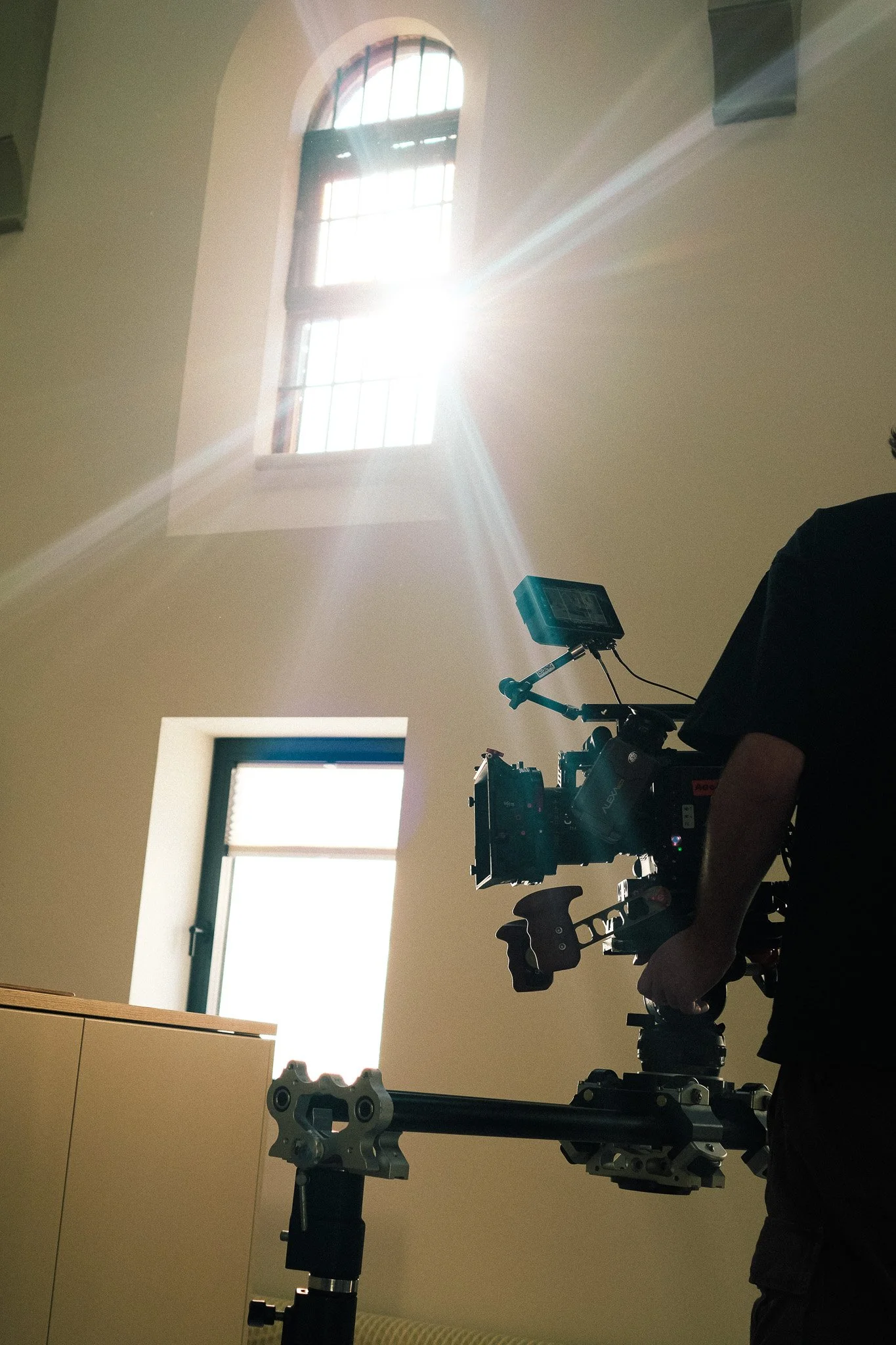 A person operating a camera on a stabilizing rig in a room with a high ceiling, two windows allowing sunlight to stream in, and a wooden cabinet in the foreground.