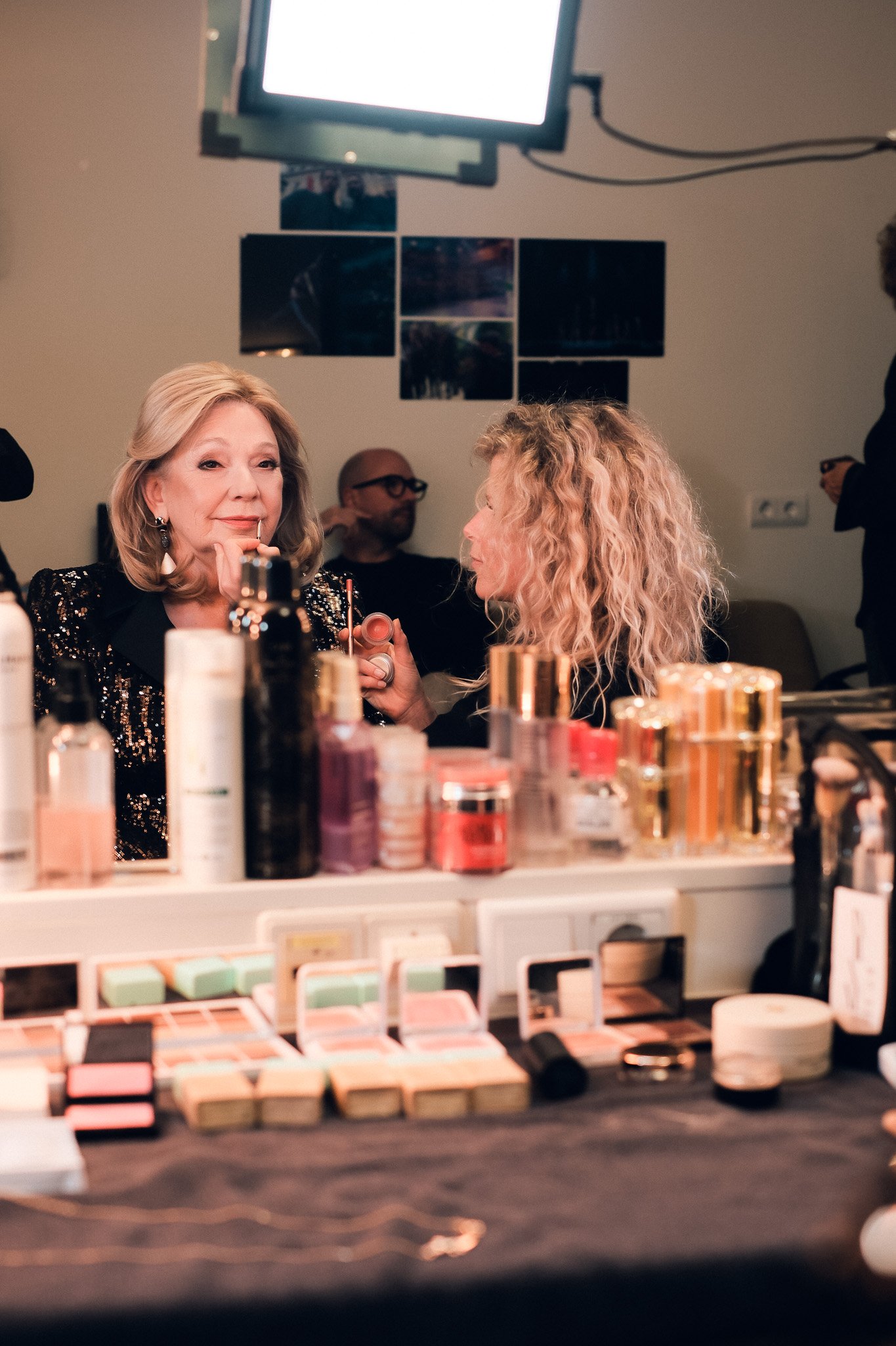 A woman with blonde hair and earrings looking into a mirror while holding makeup tools, with various makeup products on a table in the foreground, in a room with a television and photographs on the wall.