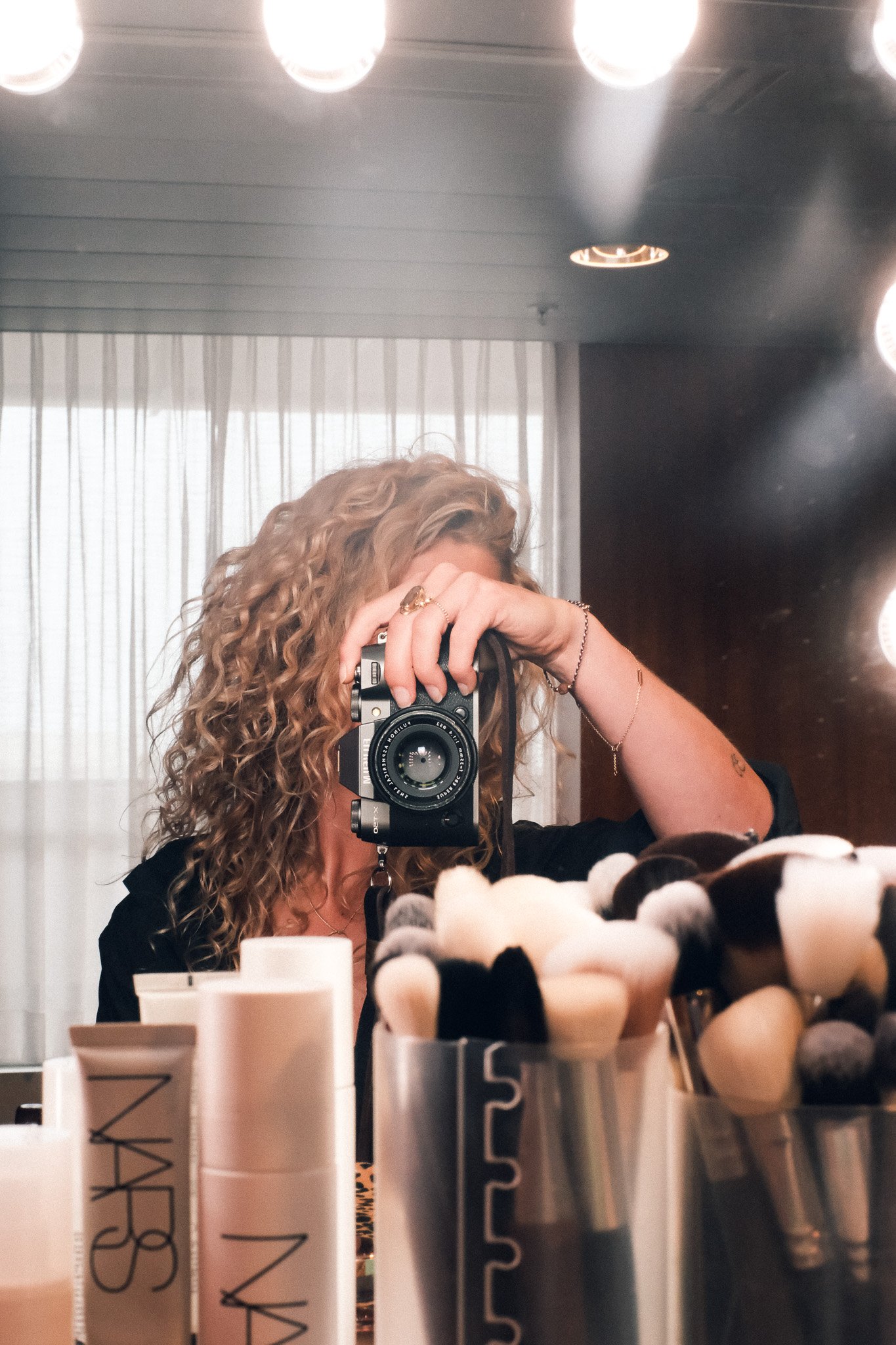 A woman with curly blond hair taking a mirror selfie in a store, with beauty products and brushes in the foreground and bright round lights on the ceiling.