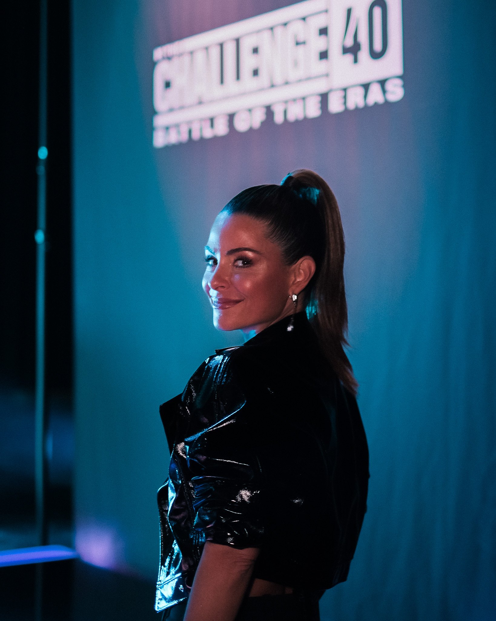 A woman with long dark hair tied in a ponytail, wearing earrings and a shiny black jacket, is looking over her shoulder and smiling in front of a blue-lit wall with a large sign that says 'SAVAGE' and some smaller text that is not fully visible.