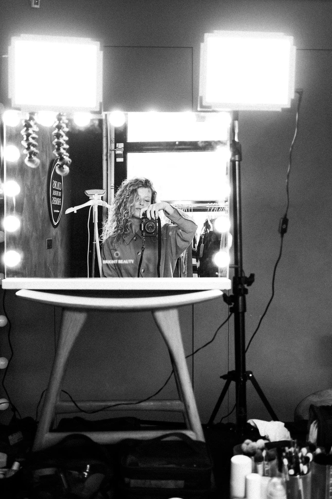 A young woman with curly hair taking a selfie in front of a mirror with bright lights, in a makeup or dressing room, with various makeup products on a table in the foreground.