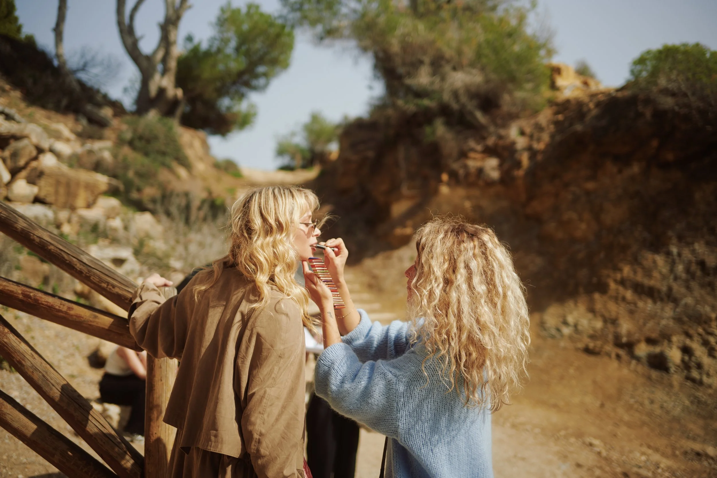 Two women with blonde, curly hair standing on a wooden bridge outdoors, one wearing a brown jacket and glasses, the other in a blue sweater, engaging in conversation amidst a natural landscape with trees and rocks.