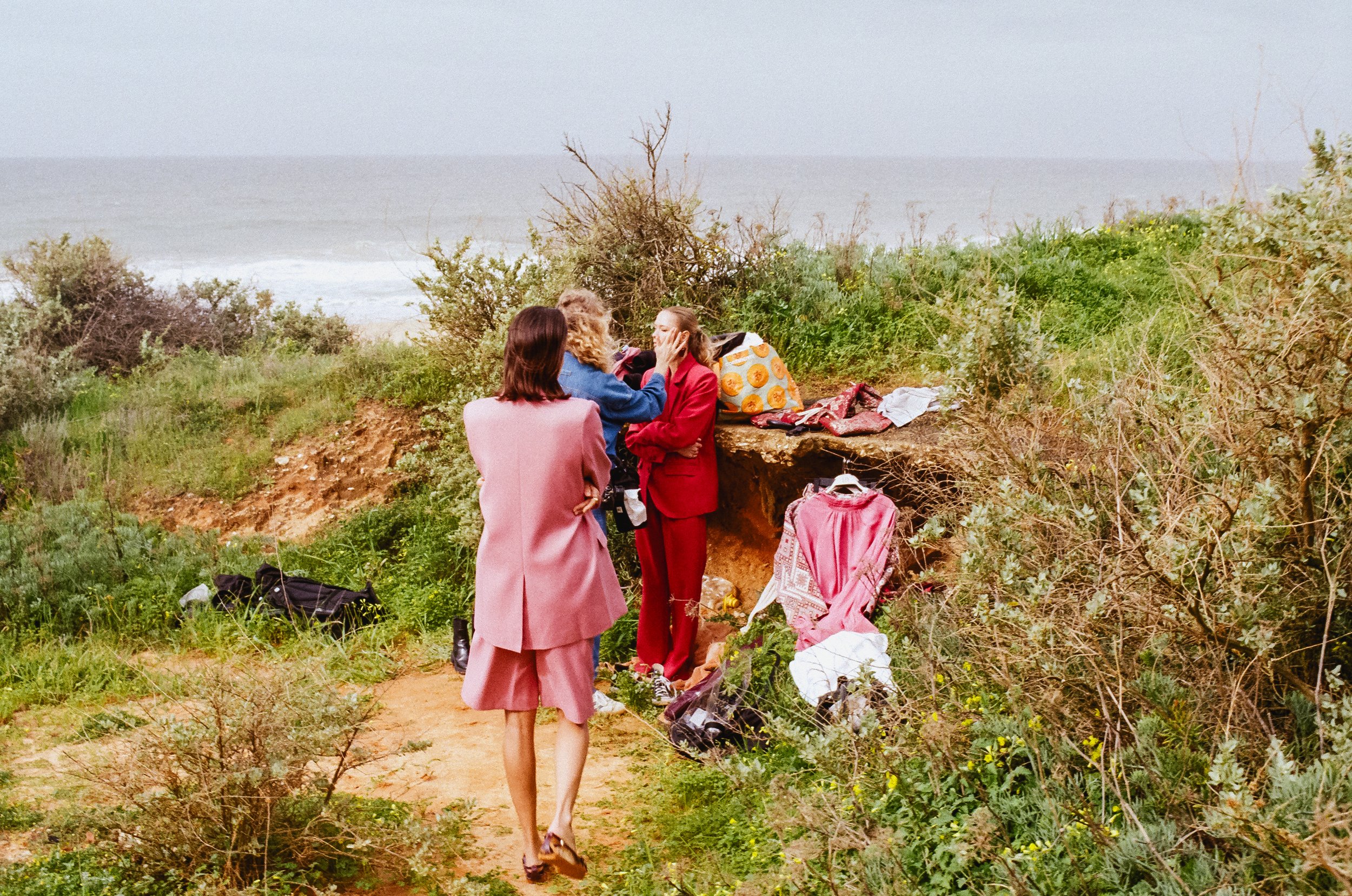 Four women standing outdoors on a dirt path near the coast, surrounded by bushes and grass, with the ocean and cloudy sky in the background, engaged in conversation.