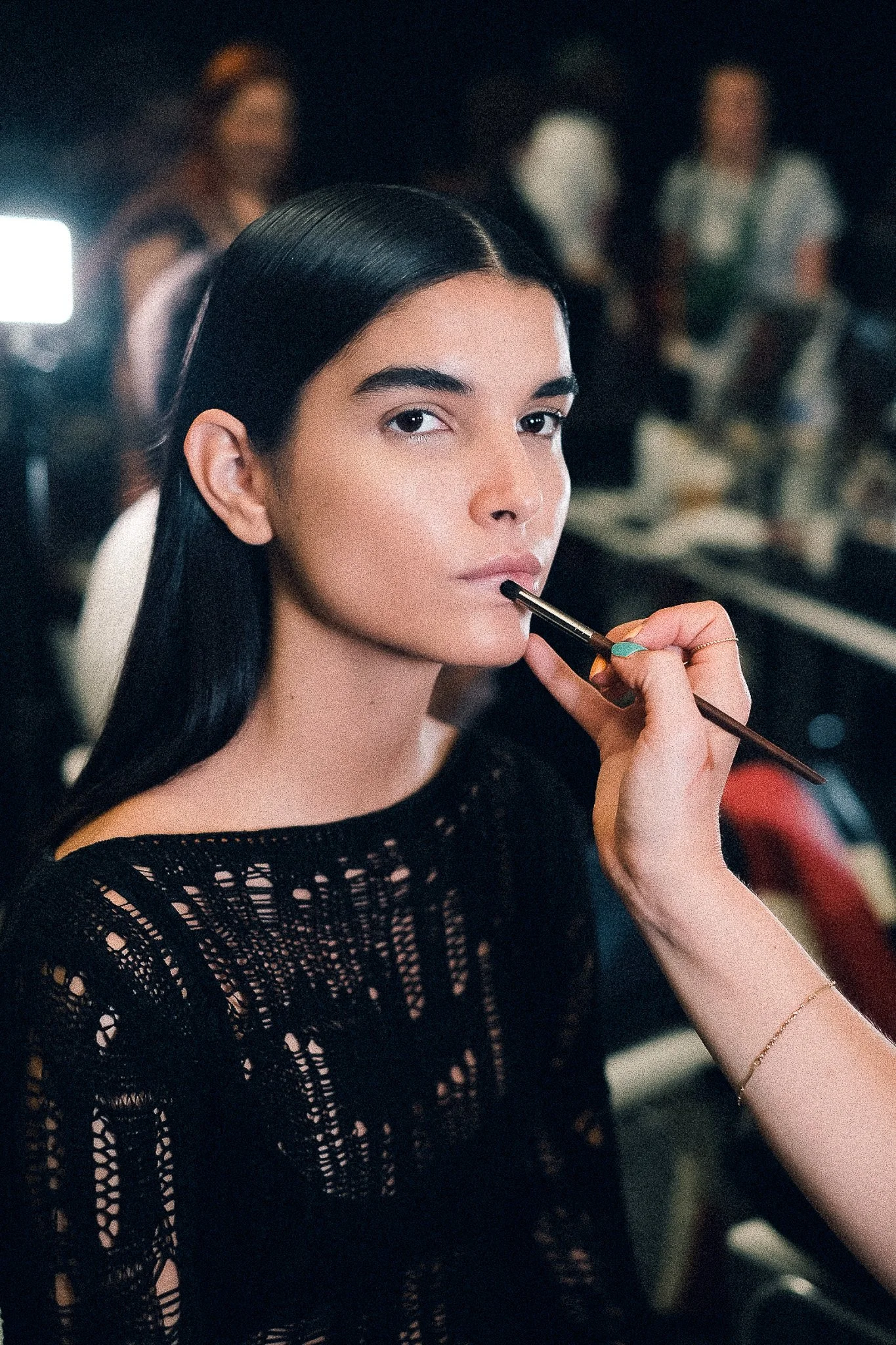 Close-up of a woman with long black hair applying makeup with a brush, wearing a black lace top, in a dimly lit room with blurred people in the background.