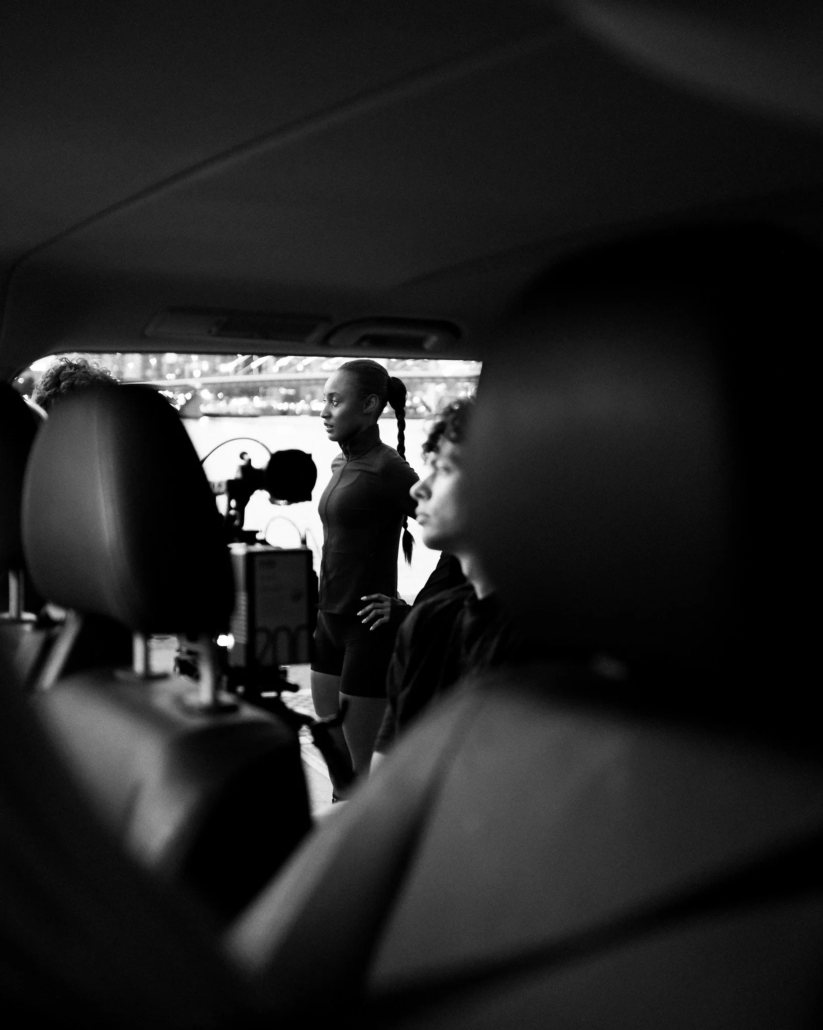 Inside a vehicle, there are people sitting in the back seat. A woman with her hair tied back is standing and looking out the window, while a person with curly hair is seated in front of her, facing forward.