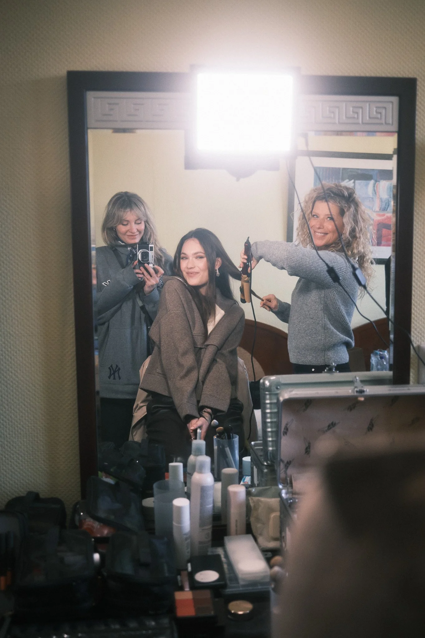 A person with straight blonde hair taking a photo of a woman with dark hair, who is sitting and smiling, in front of a mirror. Another woman with curly blonde hair is styling or blow-drying the dark-haired woman’s hair. The mirror shows a room with f