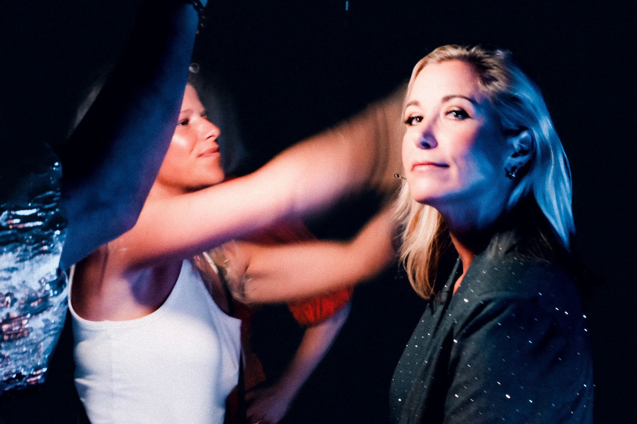 Two women dancing or posing together at a party or event, with one on the left wearing a shiny, textured top and the other on the right with blonde hair and a confident expression.