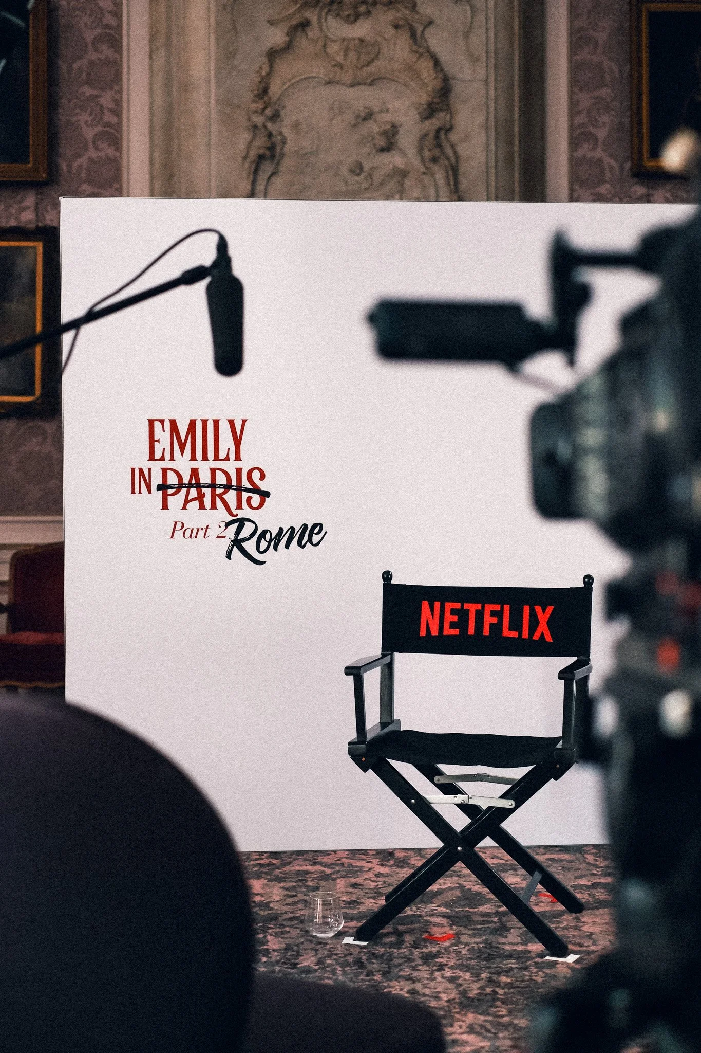 A director's chair with the Netflix logo on its backrest is centered in front of a large promotional poster for a film or series titled 'Emily in Paris Part 2 Rome'. The surroundings include ornate wall decor and a shield-shaped relief sculpture in t