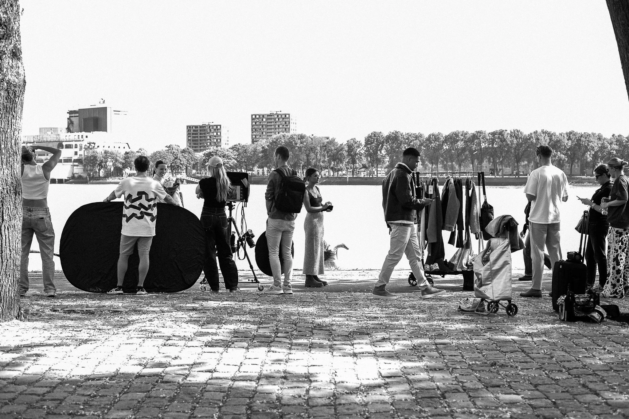 A group of people standing and socializing outdoors by a river, with trees and buildings in the background, some using cameras and phones, and clothing displayed on racks.