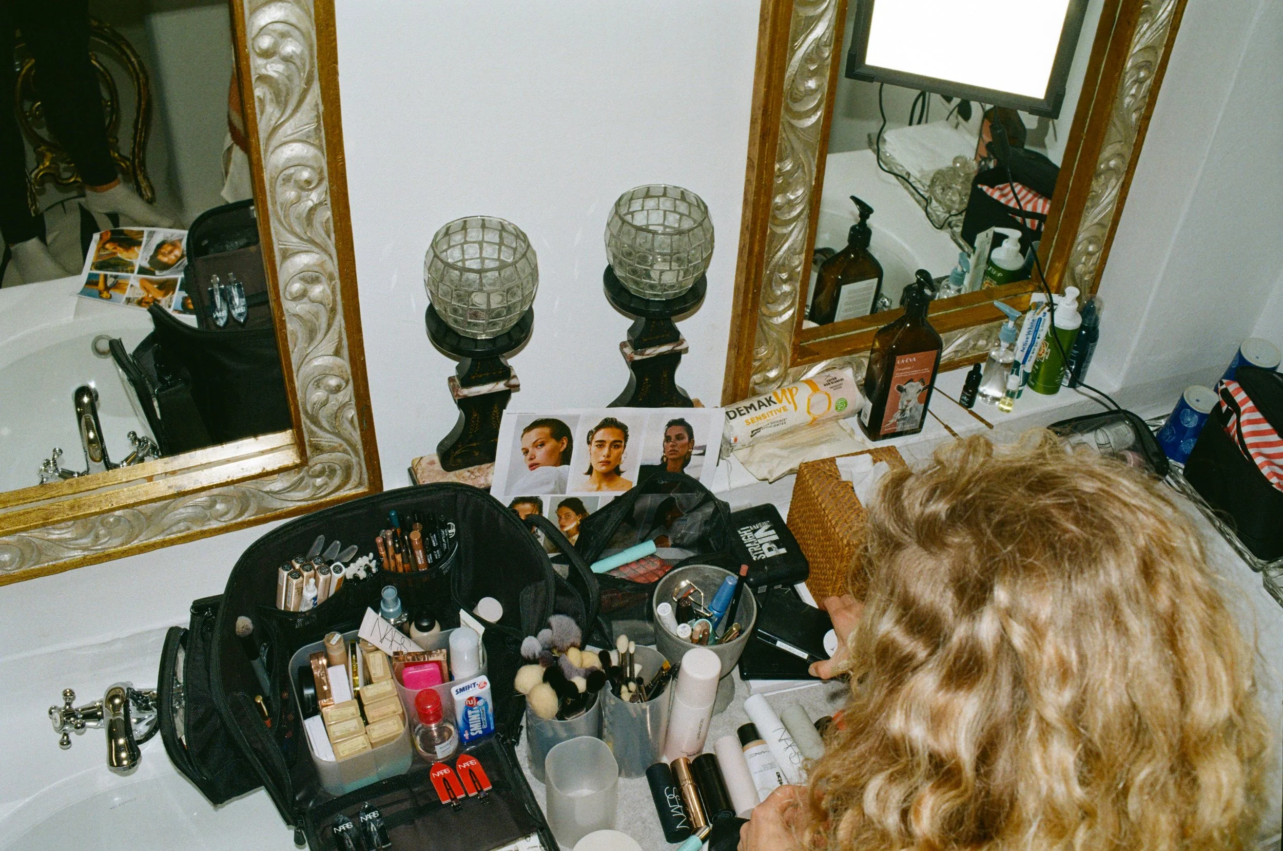 A dressing table with various makeup products and tools, a large decorated mirror, and decorative candle holders. Makeup brushes, lipstick, foundation, and other beauty products are arranged on the table. A person with curly blonde hair is seen from 