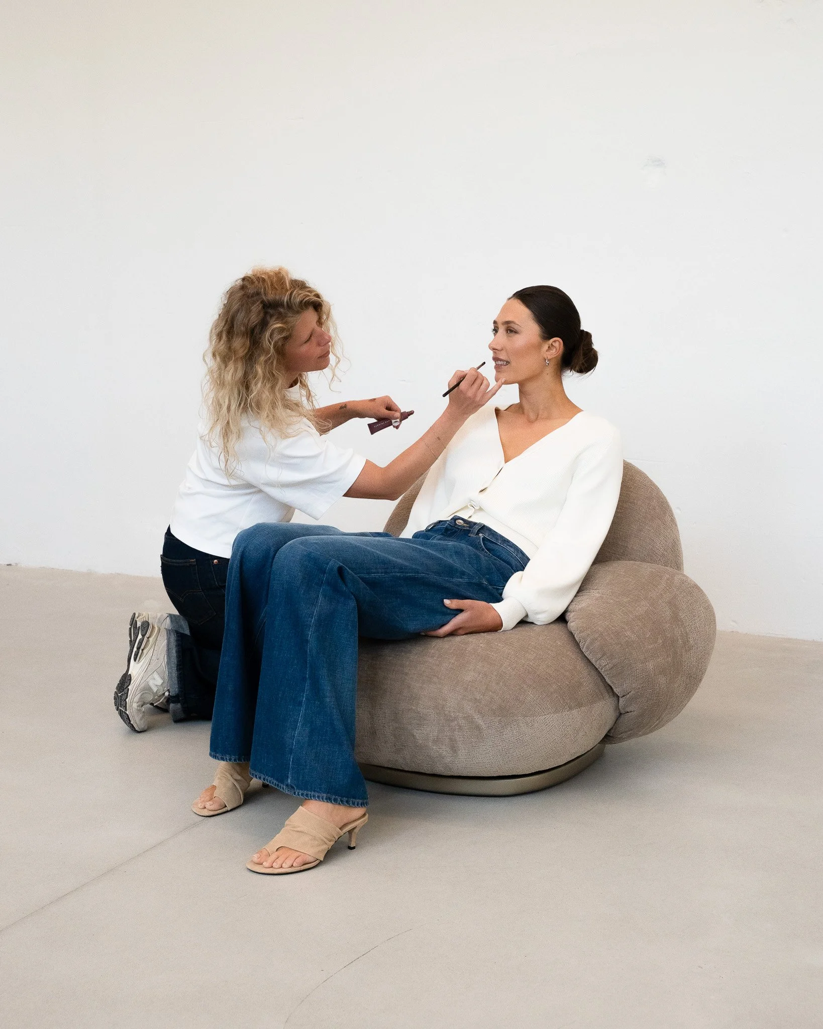 A woman is sitting on a large, rounded, cushioned chair with her legs crossed at the ankles, while another woman is applying makeup to her face. The background is plain white.