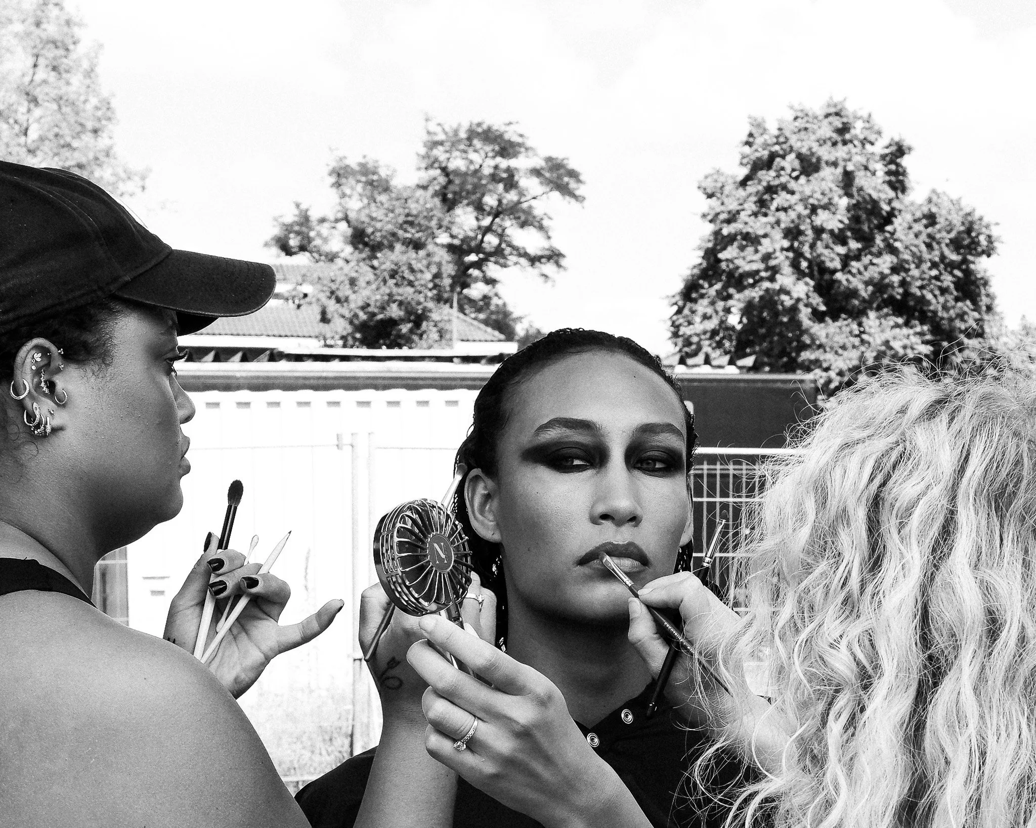 Two women at an outdoor makeup session, one applying makeup to the other, with trees and a building in the background.