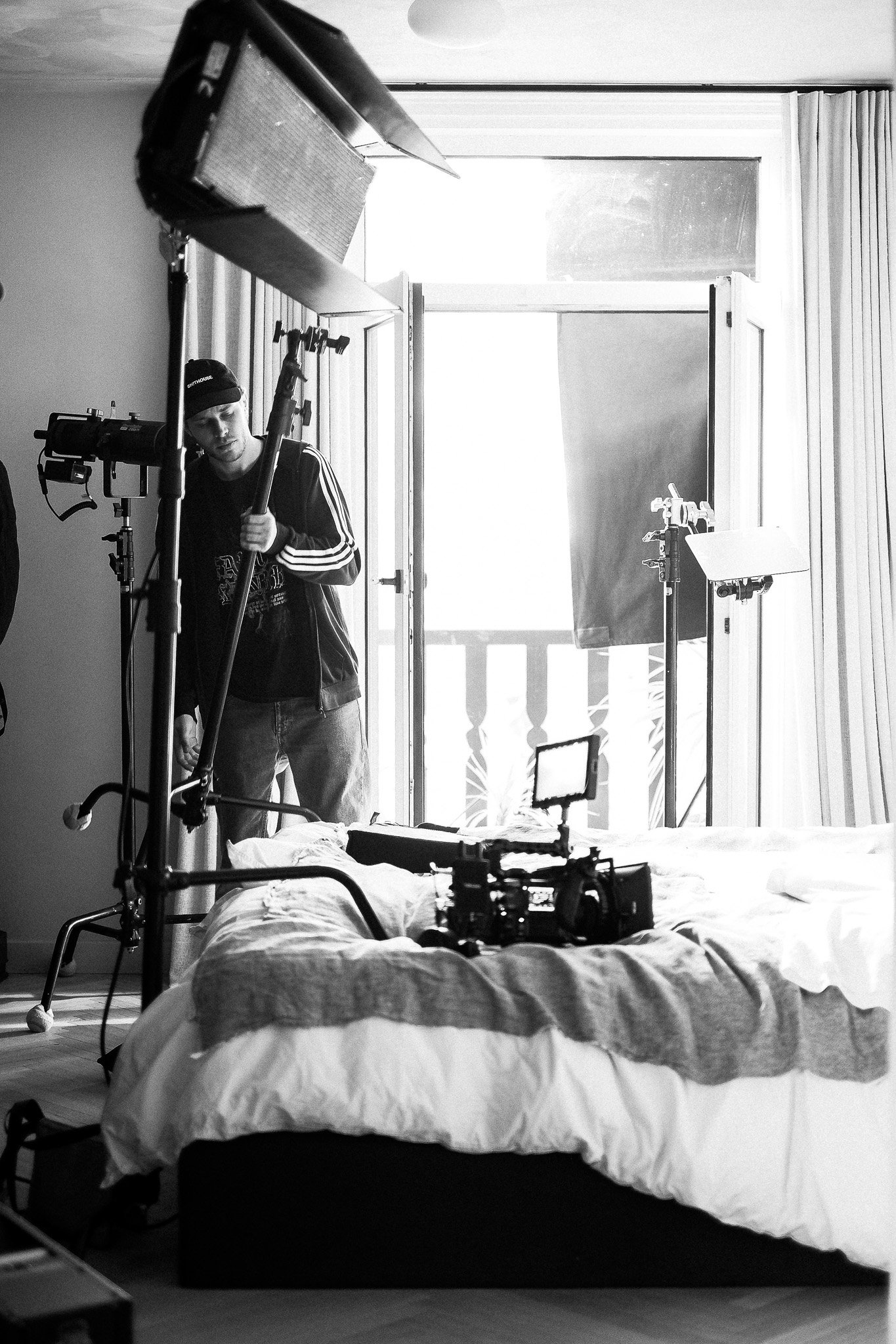 Black and white photo of a film crew in a room with large window, a person adjusting camera equipment, a bed with rumpled bedding, and various filming equipment and lighting in the foreground.
