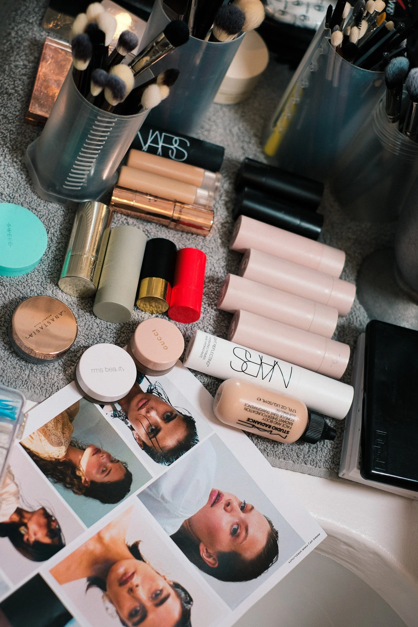 A collection of makeup brushes and cosmetic products, including lip glosses and powders, arranged on a countertop next to printed photographs of women with various hairstyles and makeup looks.