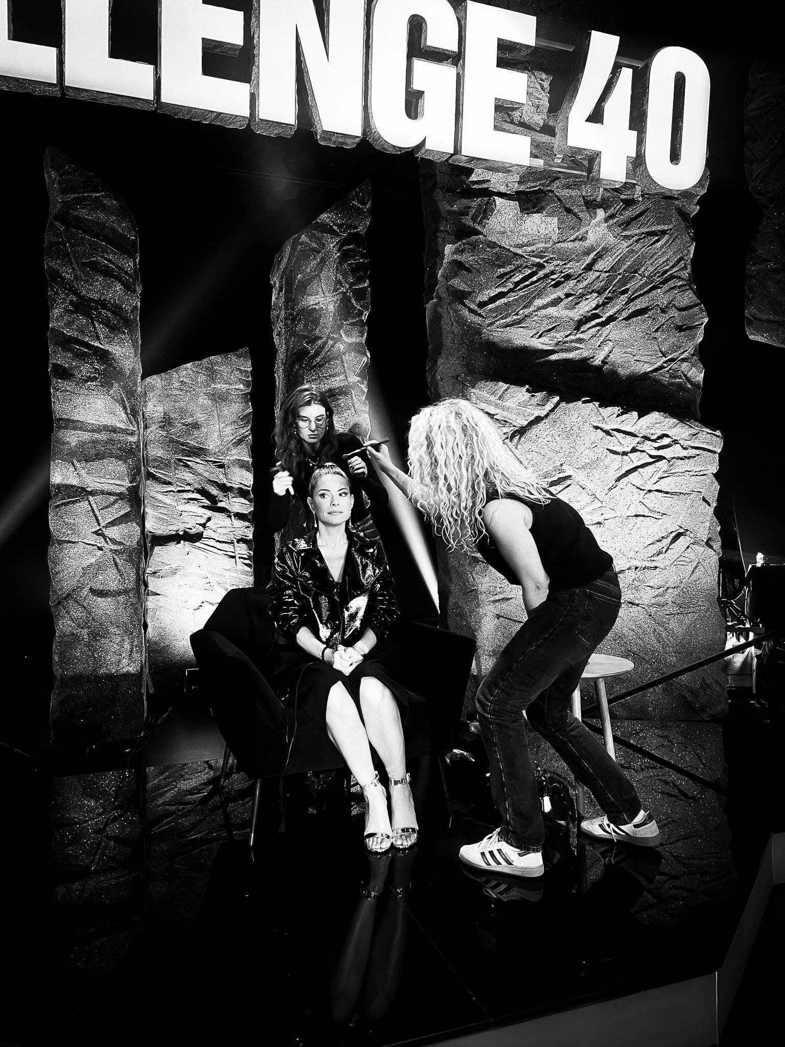 A woman in a leather jacket and high heels sitting on a chair is getting her makeup done by a makeup artist with curly hair, in front of a rock-like backdrop and large illuminated letters spelling 'LEGENE/40' hanging above.